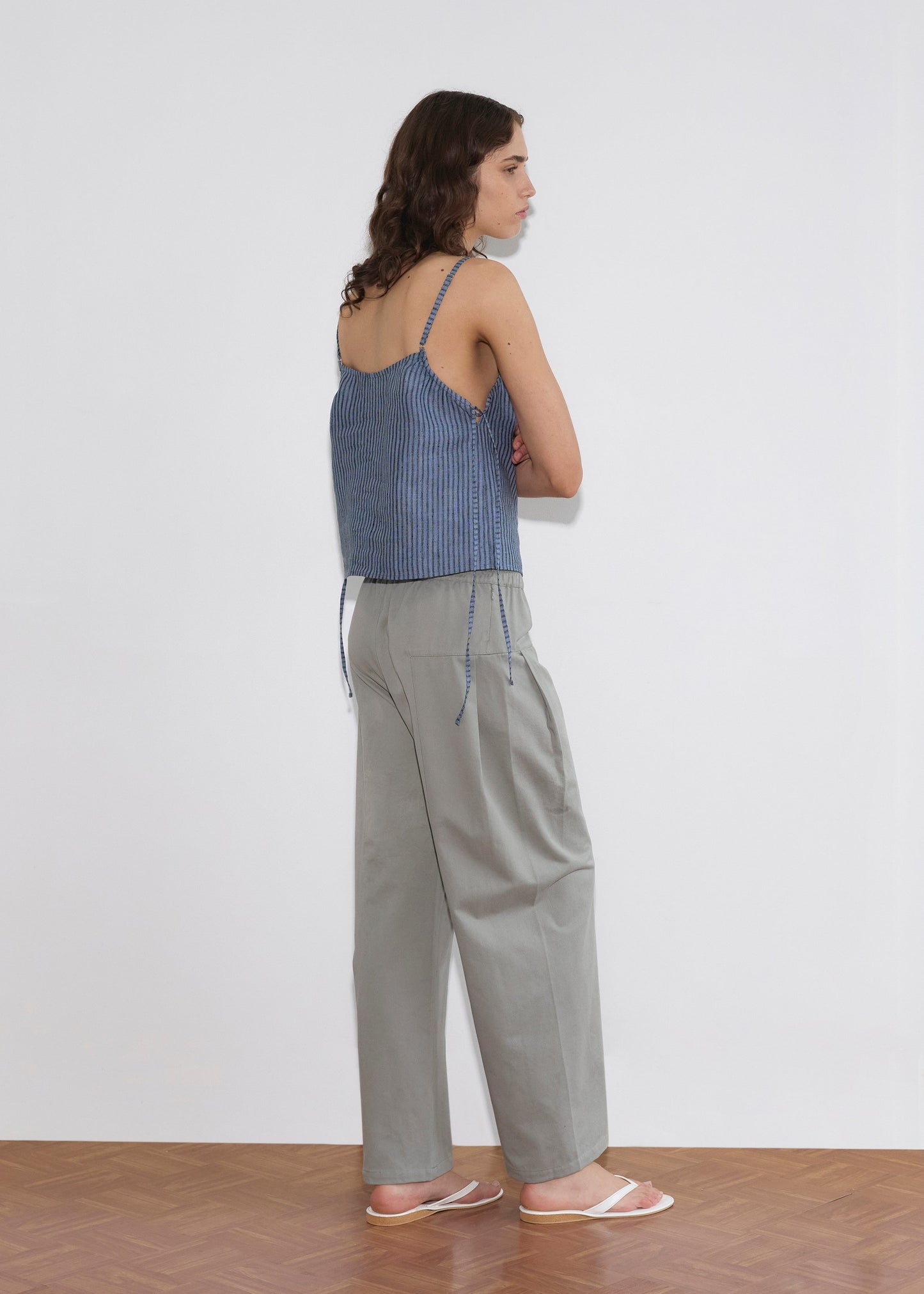 A woman with wavy brown hair stands sideways against a white wall, wearing a blue striped sleeveless tie-strap top, Pleat Pant - Sage Grey by Deiji Studios, and white sandals on a wooden floor.