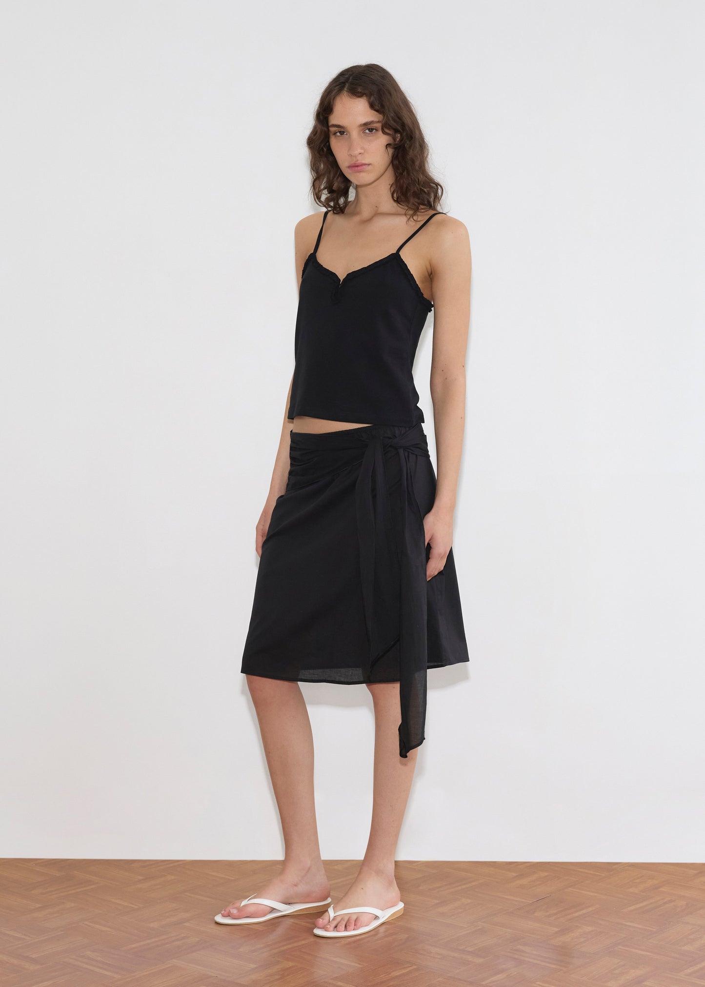 A person wearing the Clasp Singlet - Black by Deiji Studios, a black tie-waist wrap skirt, and white flip-flops stands in front of a plain white wall on a wooden floor.