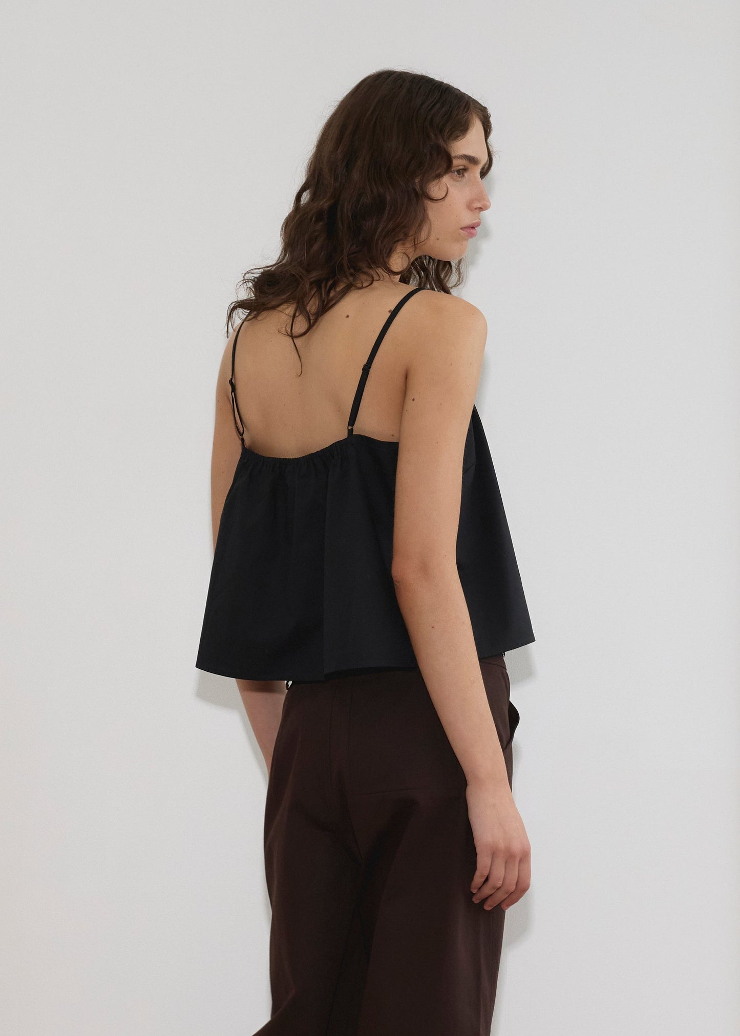A person with wavy brown hair stands sideways against a plain light background wearing the Double Layer Top - Black by Deiji Studios, styled loosely and paired with dark brown pants.