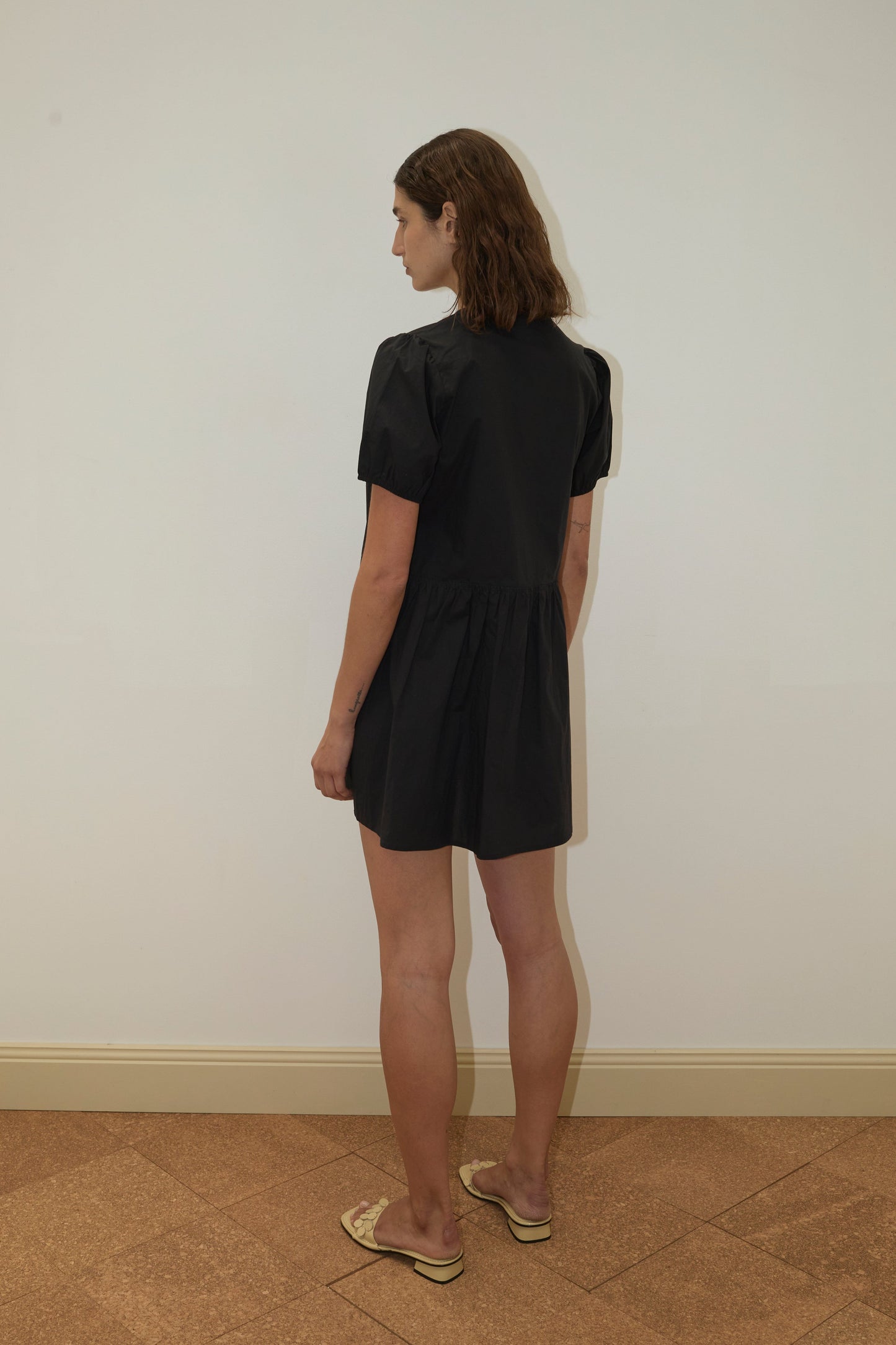 A person with shoulder-length brown hair stands sideways against a white wall and brown tiled floor, wearing the Pocket Mini Dress - Black by Deiji Studios with short sleeves and beige sandals.