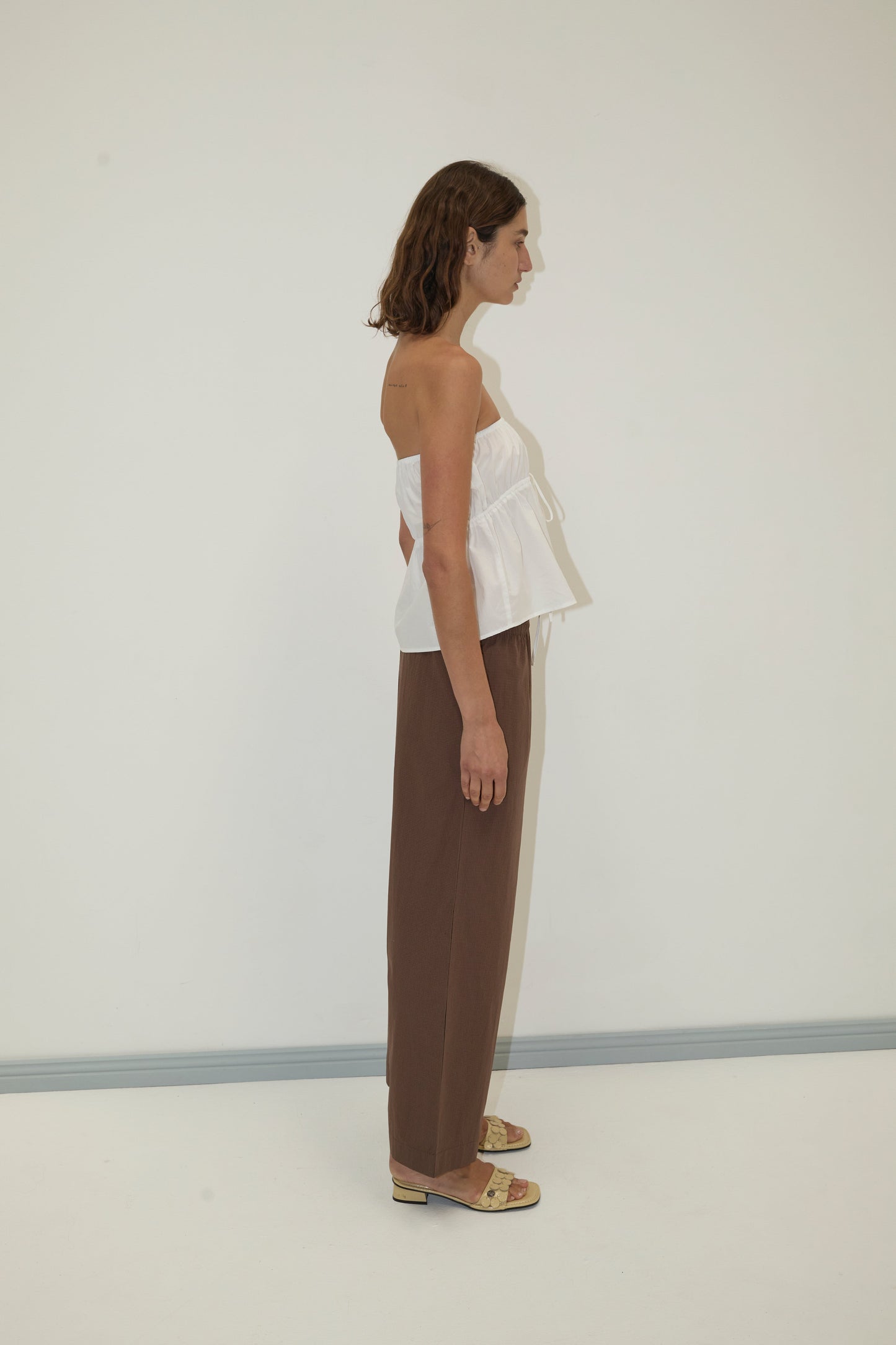 A woman with shoulder-length brown hair stands in profile against a plain white wall, wearing a strapless cotton top - white from Deiji Studios, loose brown pants, and beige sandals.
