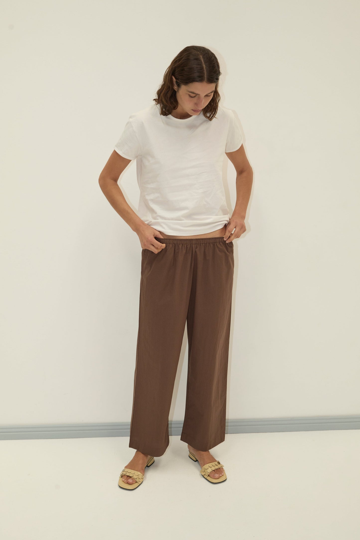 A person stands against a plain white wall, wearing a white t-shirt, the Ease Trouser - Carob Check with an elastic waist, and open-toed sandals, looking down with hands in their pockets.