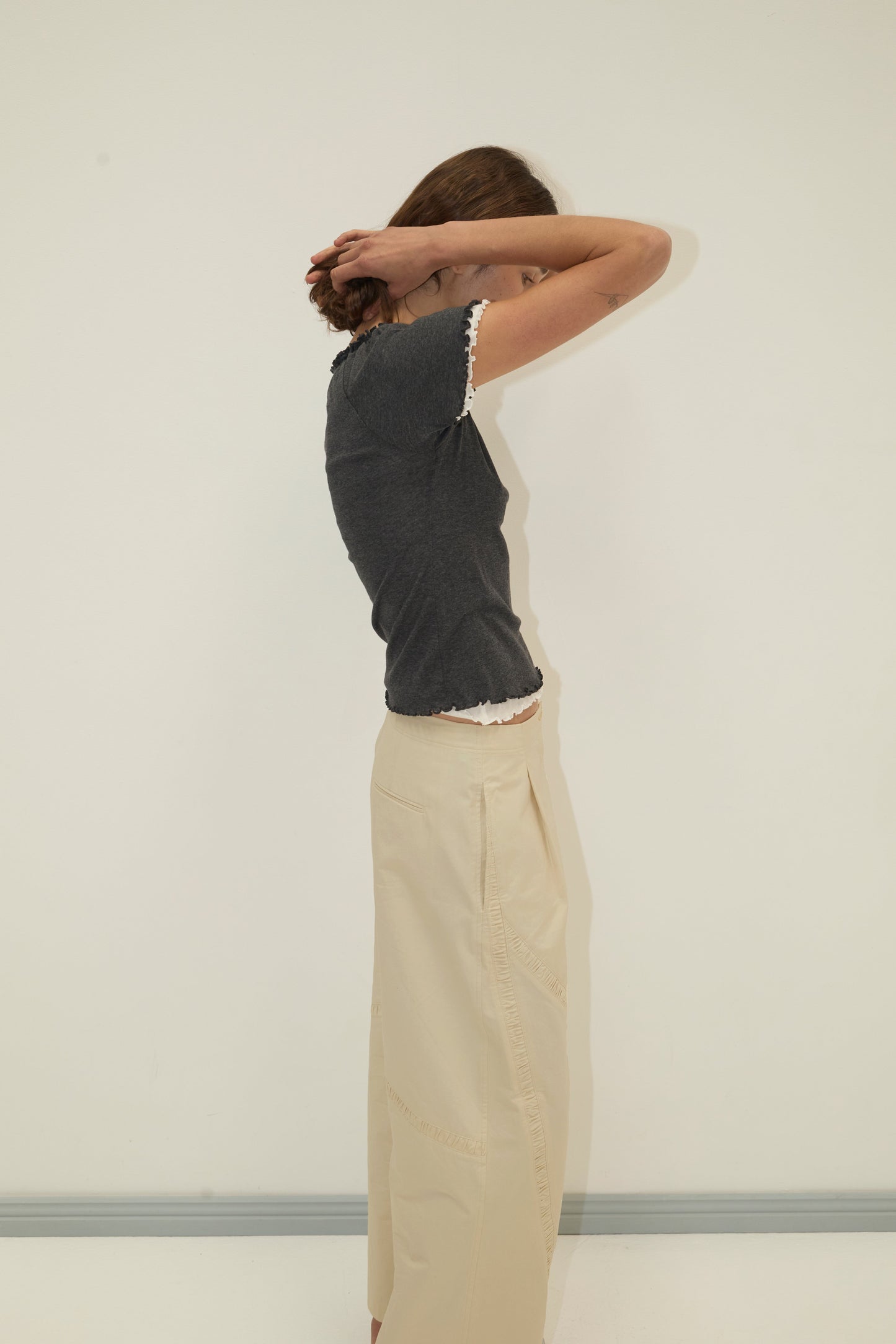 A person stands sideways against a plain light wall, lifting their arms to adjust their hair. They wear the Deiji Studios Channel Pant in Stone and a dark short-sleeve top; their face is partially obscured.