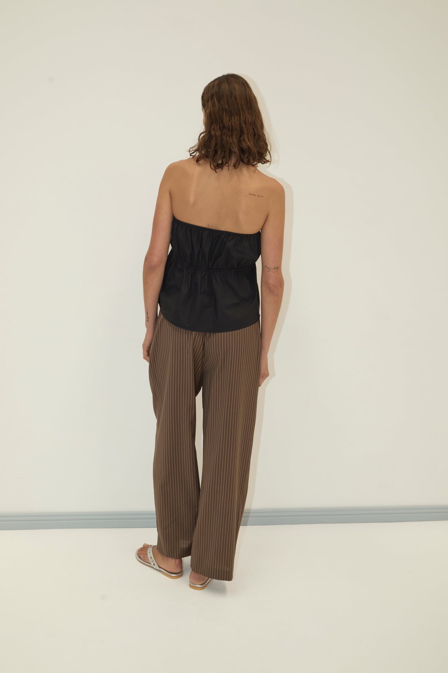 A person with shoulder-length brown hair faces a plain white wall, wearing a black strapless top, Pure Wool Ease Trouser - Sable Stripe by Deiji Studios, and sandals. A tattoo is visible on their upper back.