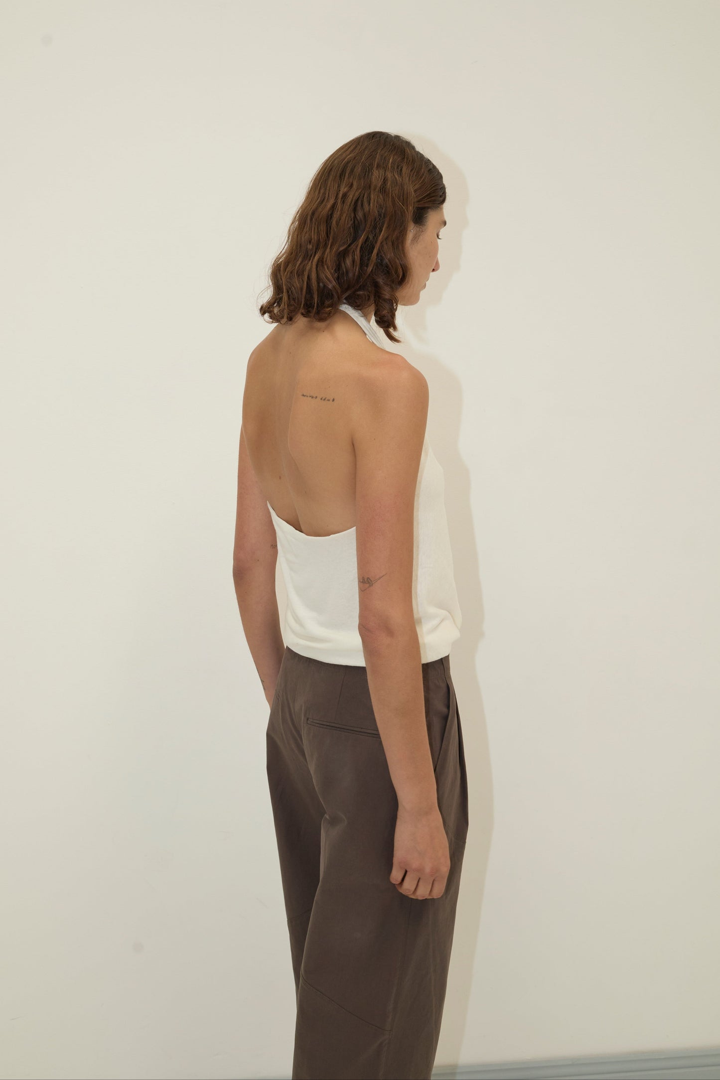 A person with shoulder-length brown hair wears a white halter top and the Flag Tie Pant in Dark Brown, standing sideways against a plain white wall with arms relaxed and looking down.