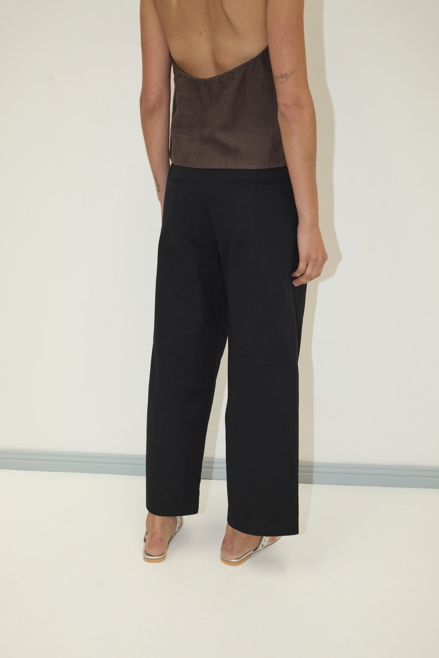 A person faces away from the camera in a brown strapless top, Flag Tie Pant - Black by Deiji Studios, and clear-heeled shoes. The background is plain white with a gray baseboard.