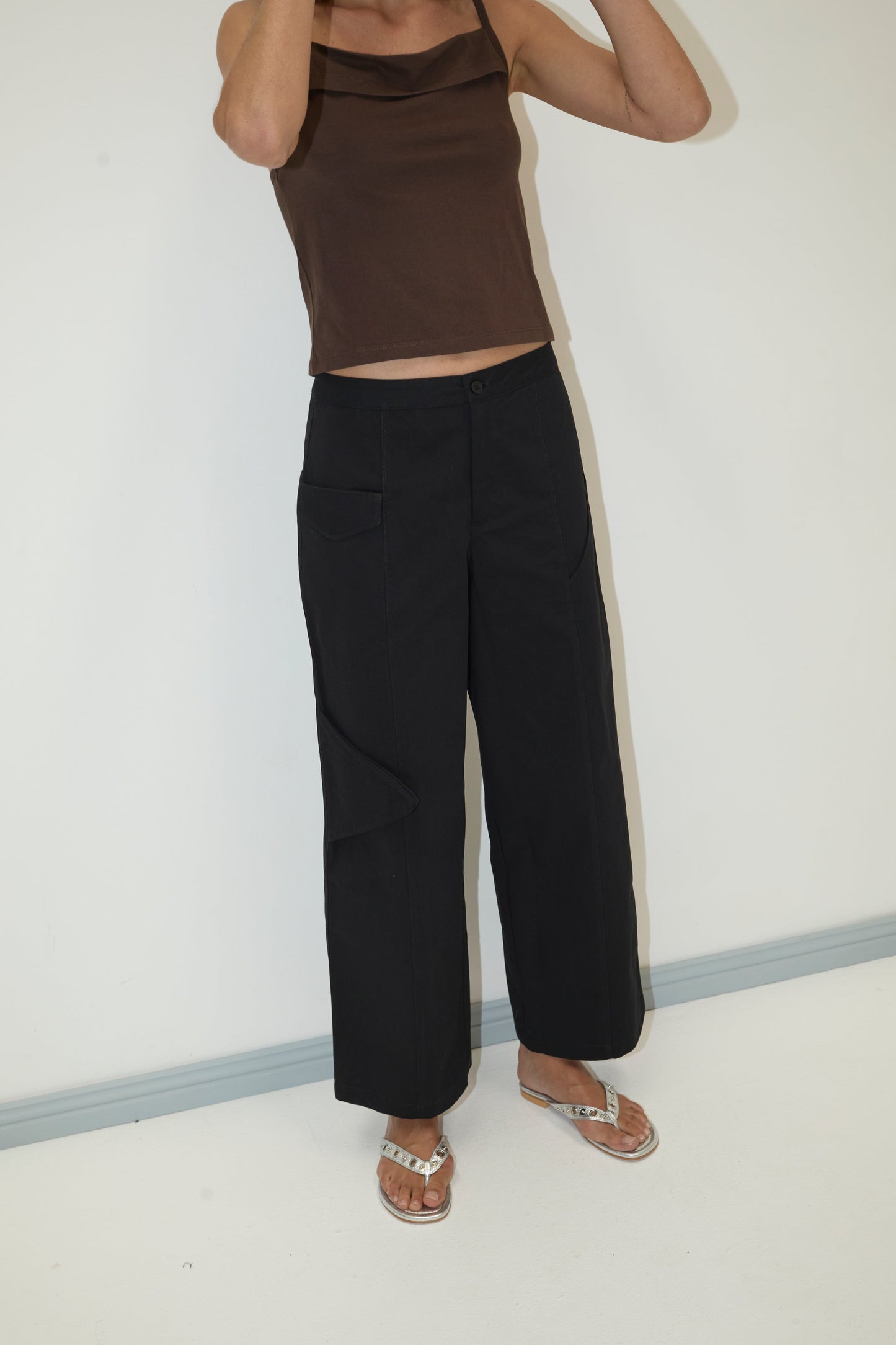 A person in a brown sleeveless top, Pocket Pant - Black by Deiji Studios, and silver sandals stands against a plain white wall. Their head is cropped out of the frame.