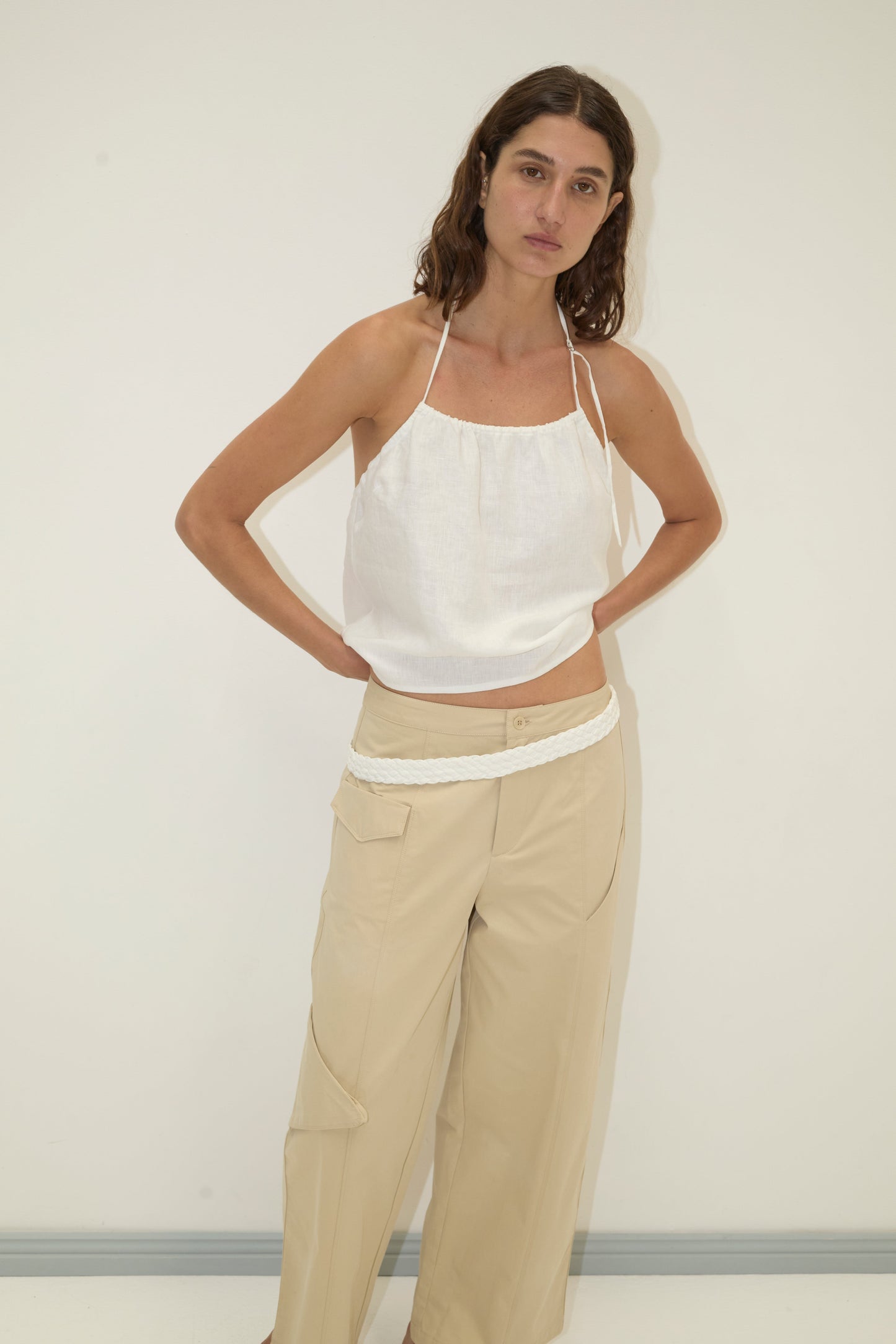 A woman stands against a white wall, wearing a white sleeveless halter top and Deiji Studios’ Pocket Pant - Stone in organic cotton with side pockets and a white braided belt, her hands resting on her hips.