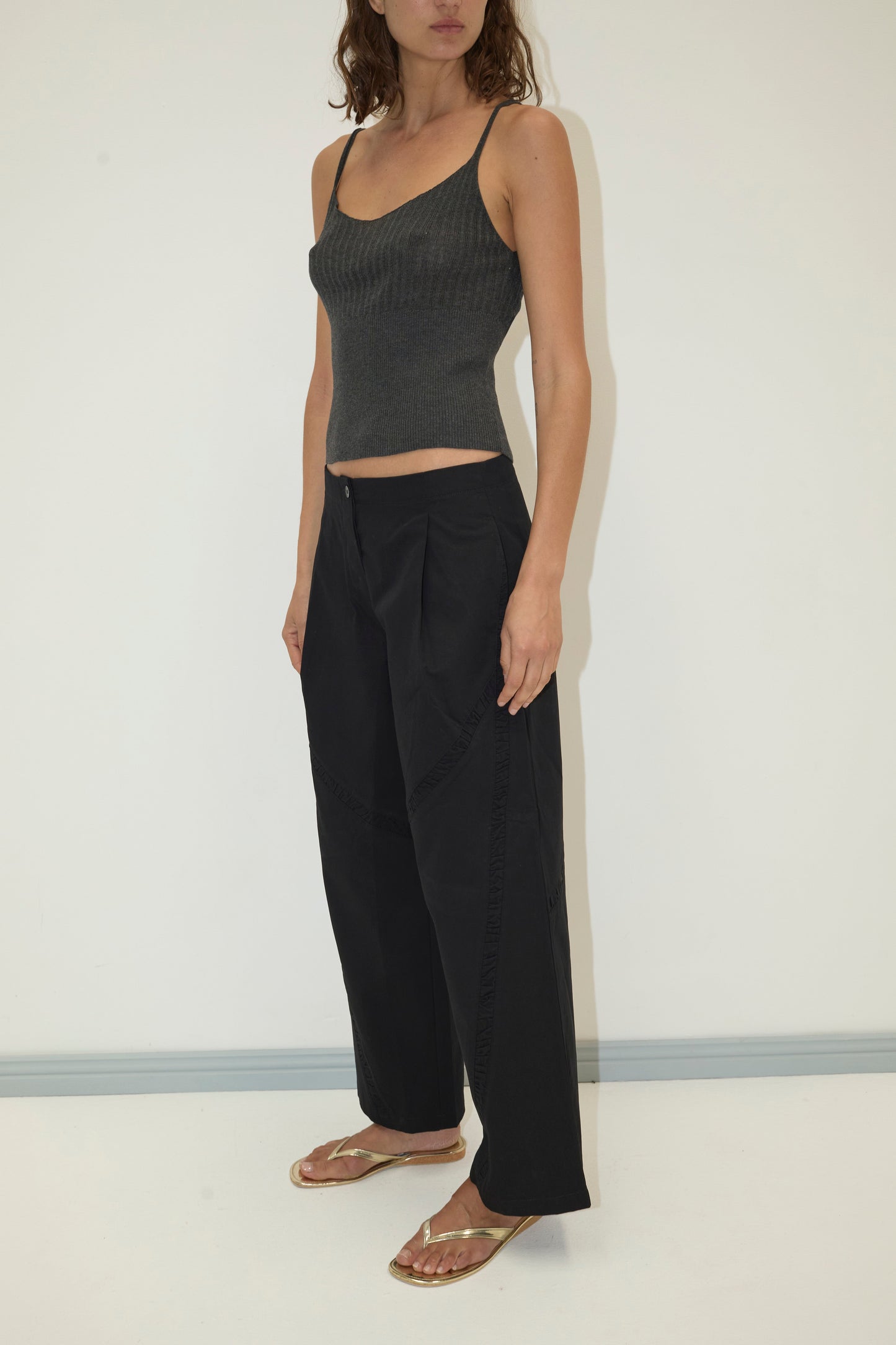 A person stands against a plain background, wearing a fitted dark gray spaghetti strap tank top, the Channel Pant - Black by Deiji Studios, and gold flip-flops. Their arms rest at their sides and their face is partially out of view.