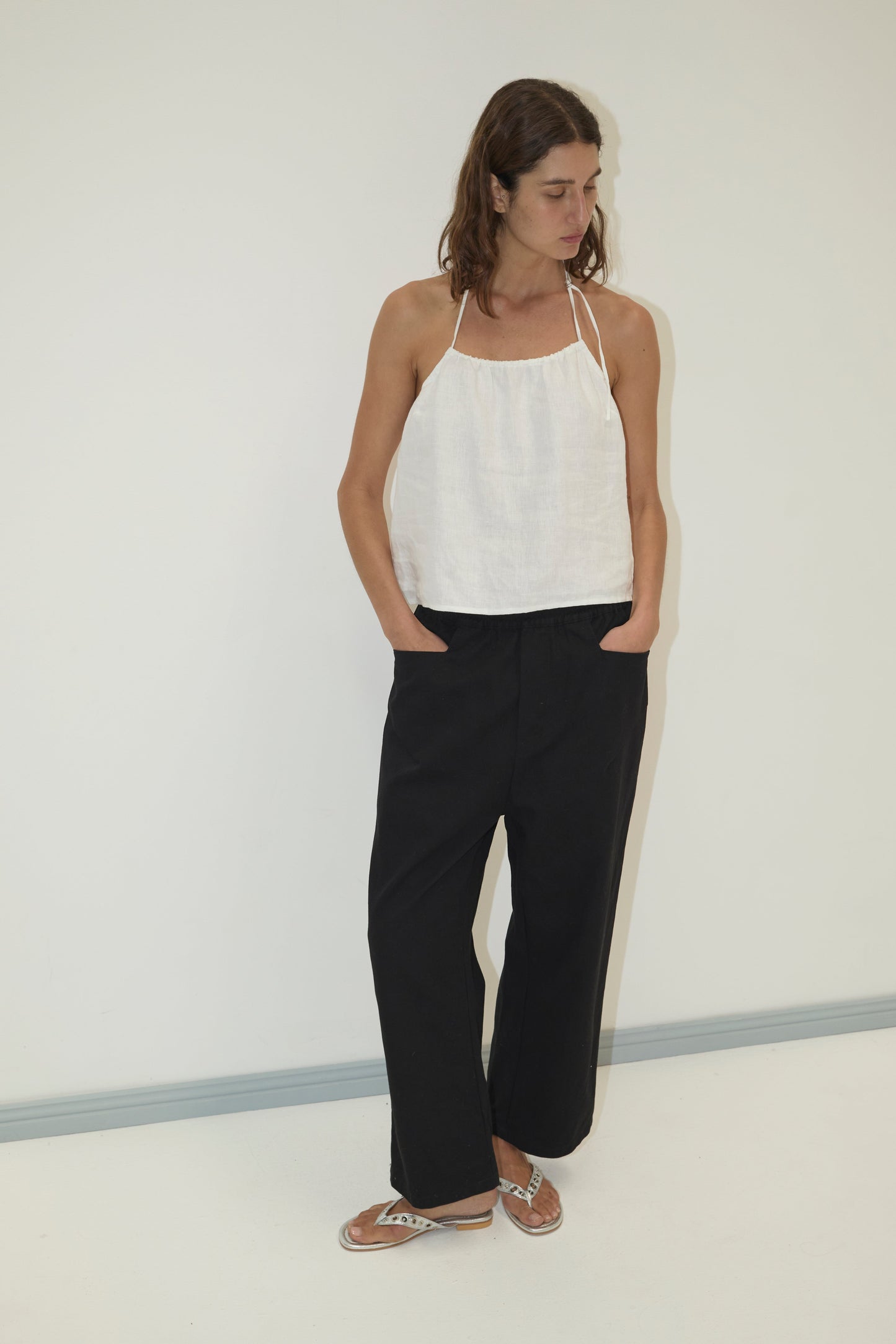 A woman stands against a plain white wall, wearing a sleeveless white halter top, the Square Pocket Pant in black, and brown sandals. She has shoulder-length brown hair, hands in pockets, and looks down to her left.