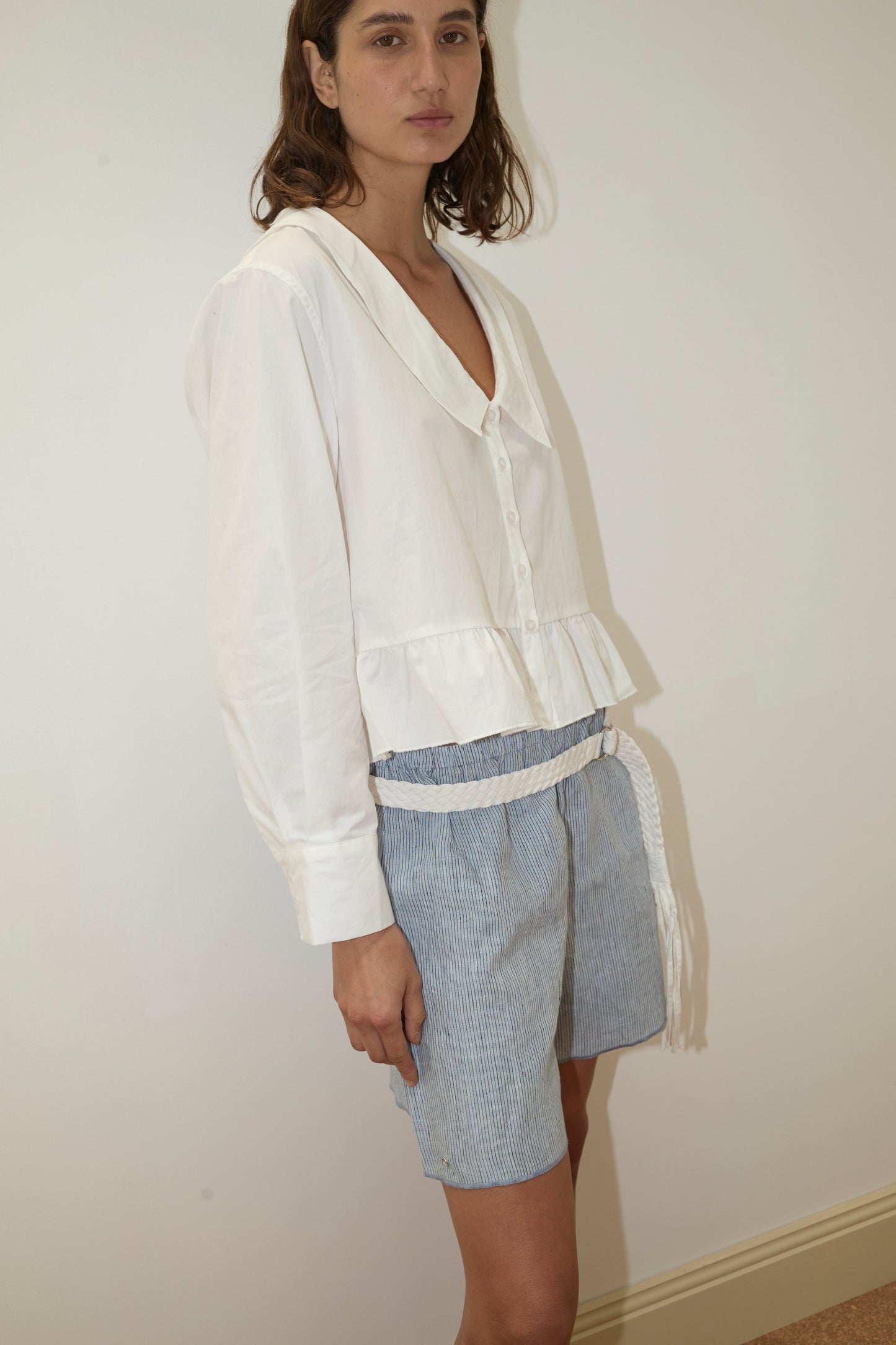 A person stands indoors against a plain wall, wearing the white Sweep Shirt from Deiji Studios paired with blue-and-white striped shorts featuring a white tasseled belt.