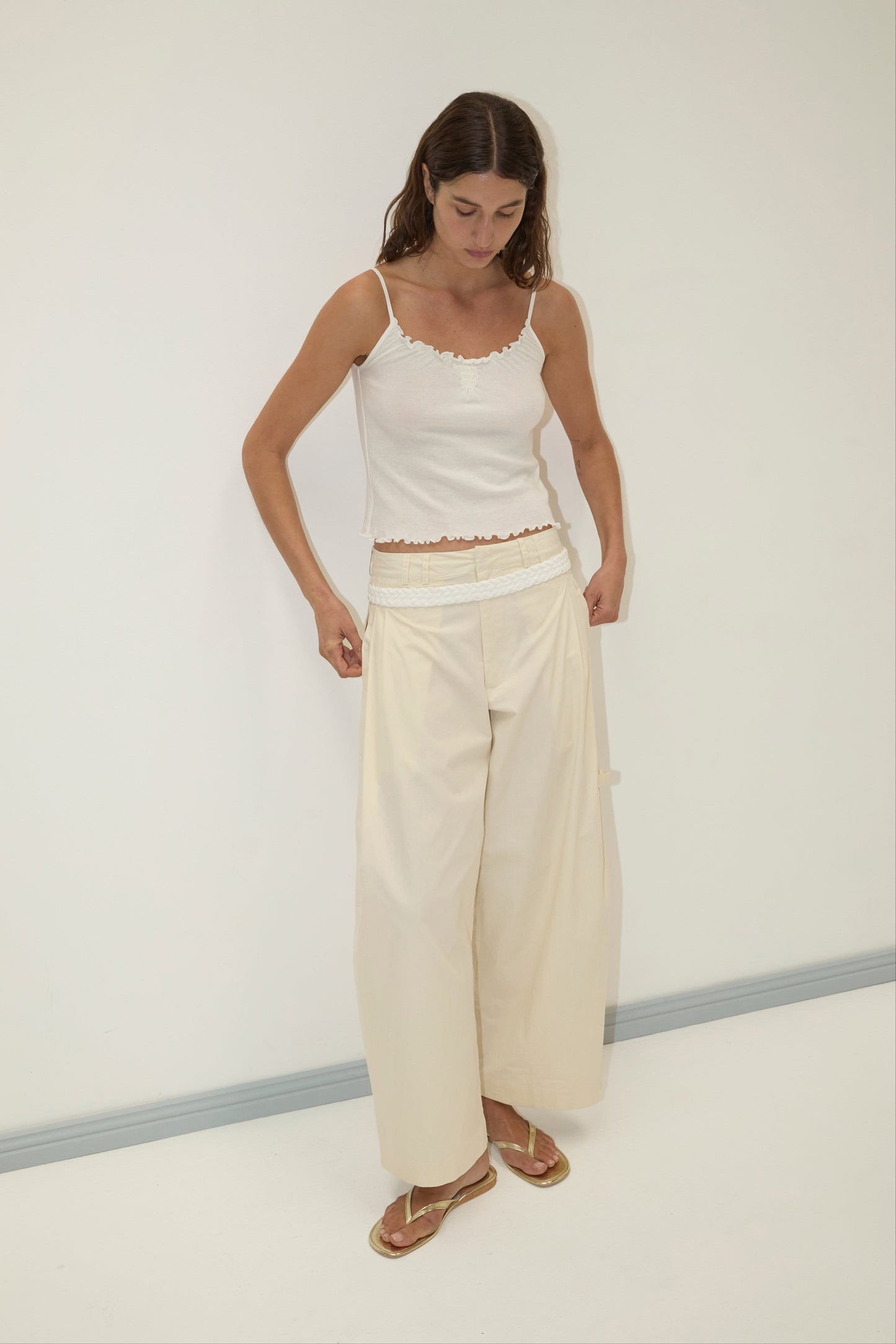 A woman in a minimal room wears a white tank, the Deiji Studios Tab Pant - Off White (relaxed, wide leg), and sandals. She looks down as she adjusts her pants against a plain, light-colored background.