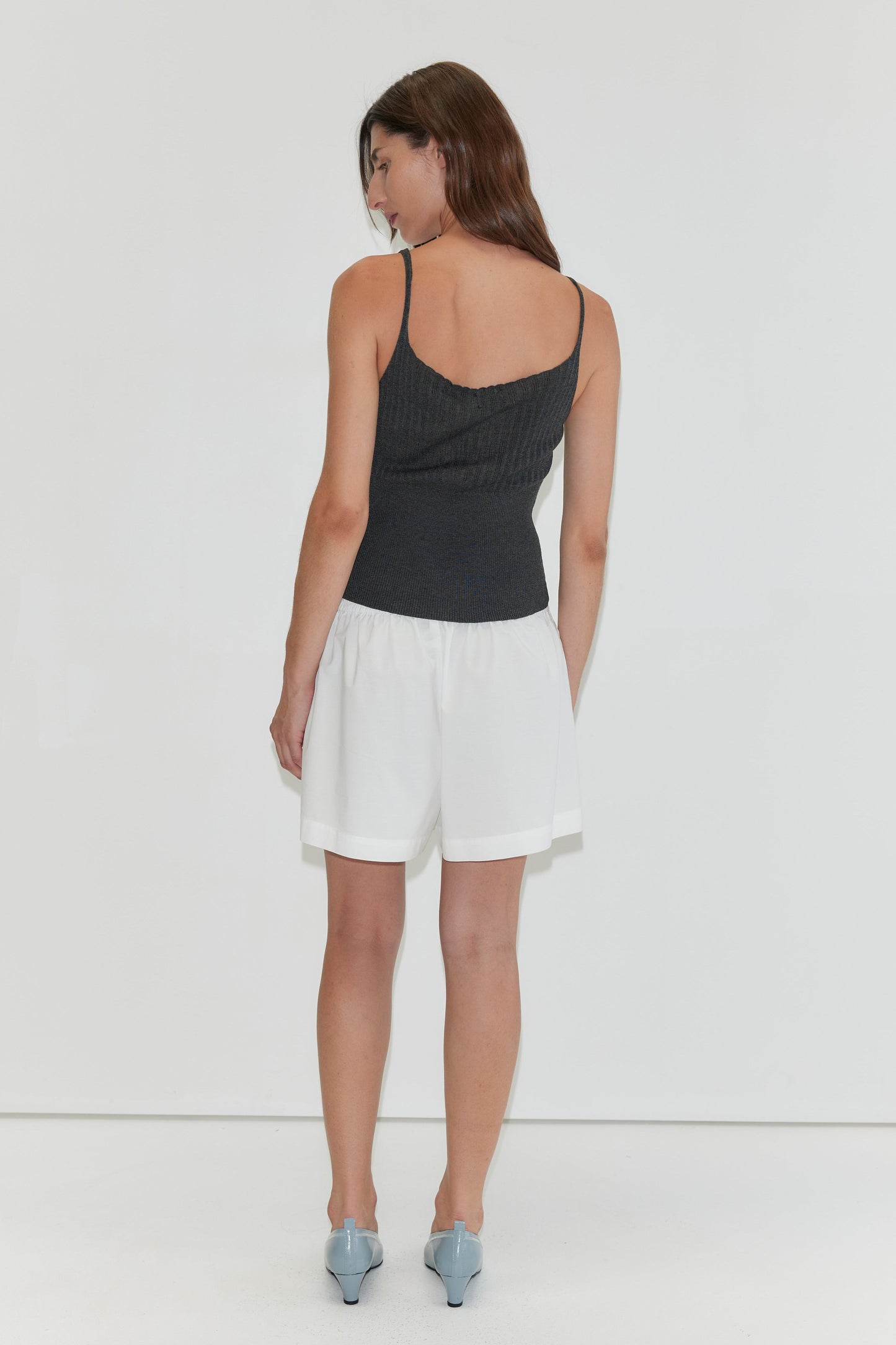 A woman faces away in a dark sleeveless ribbed top, Organic Cotton Boxer - White from Deiji Studios with an elastic waist, and light blue heeled shoes, set against a plain white background.