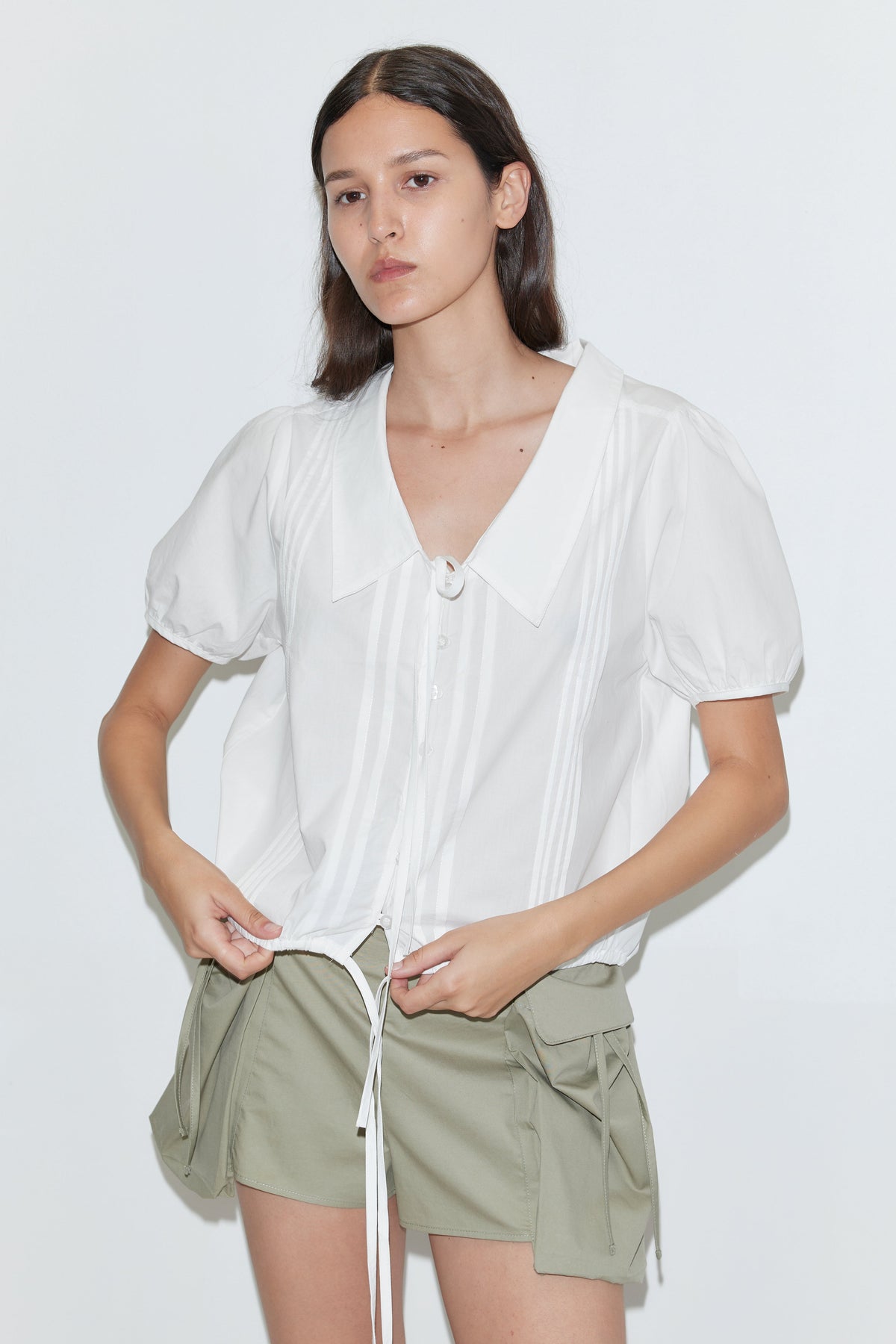 A person wearing the Deiji Studios Fine Collar Top in white cotton poplin with a wide collared v-neck and green shorts with pockets stands against a plain white background.