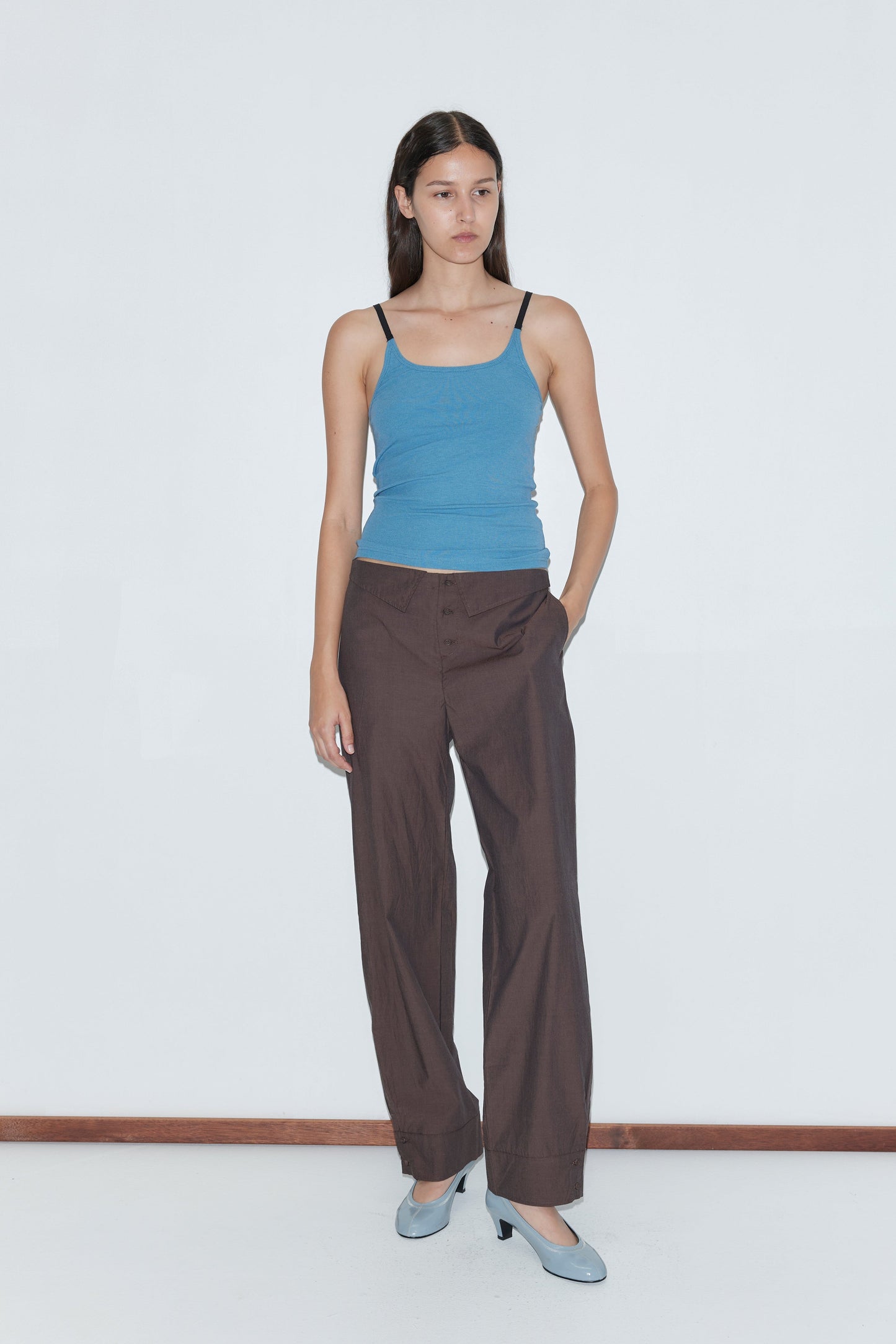 A woman stands against a plain white wall wearing a blue tank top, Collar Point Pant in Toast by Deiji Studios, and light blue heels. One hand is in her pocket and she has a neutral expression.