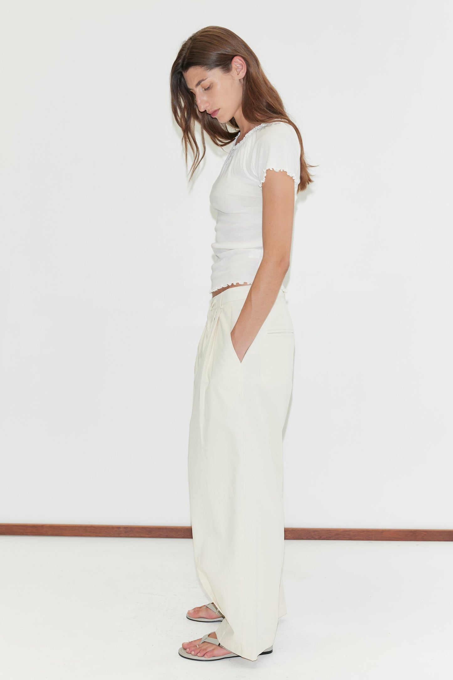 A woman with long brown hair stands in profile against a white background, wearing a short-sleeve white top, Deiji Studios Turn Pant in off white with an adjustable drawcord, and sandals; her hands are in her pockets and head slightly bowed.
