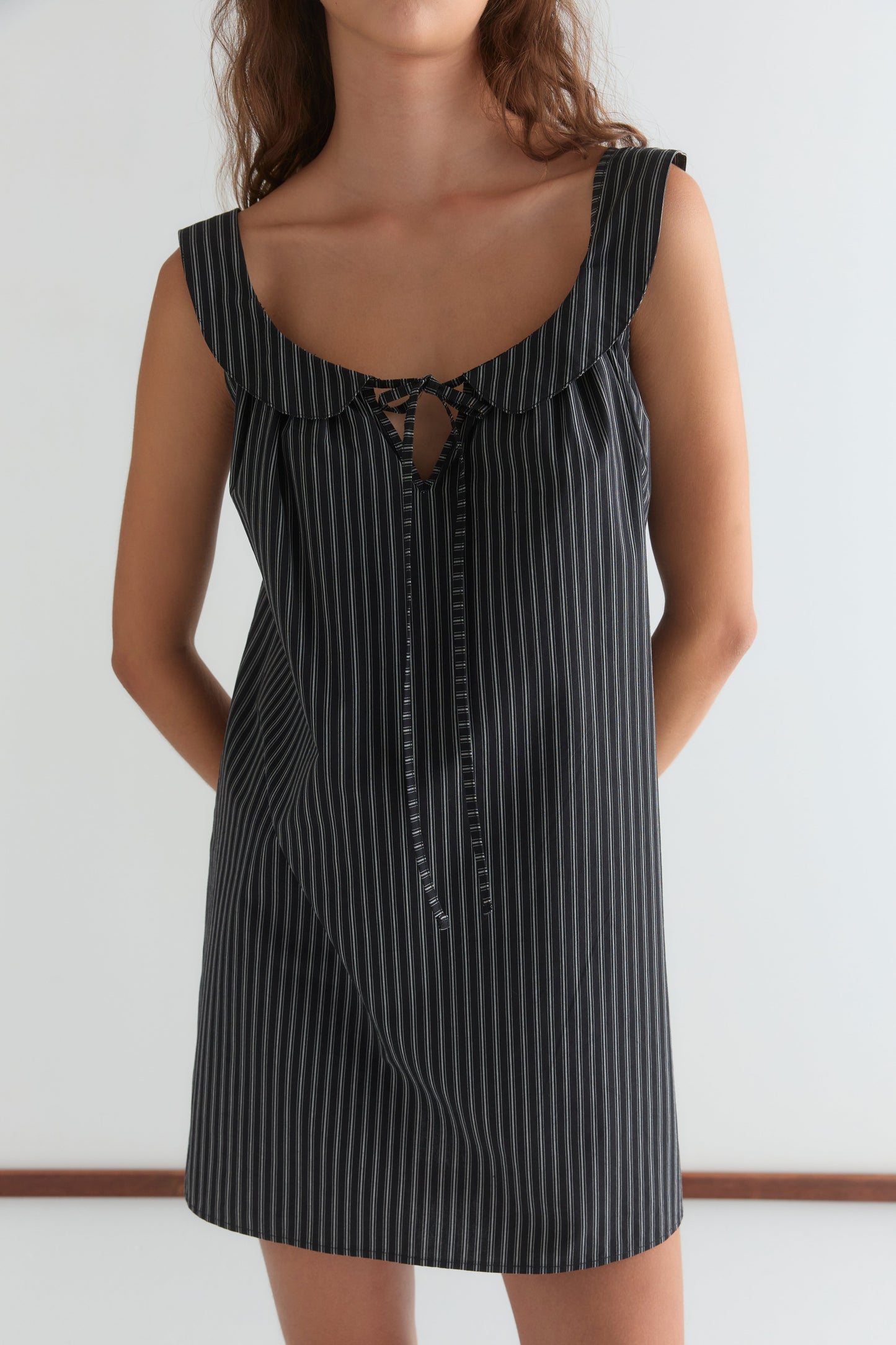 A woman wears the Deiji Studios Rounded Neck Dress— a short, black, sleeveless piece with thin white stripes, ruffled neckline, and a small front tie.