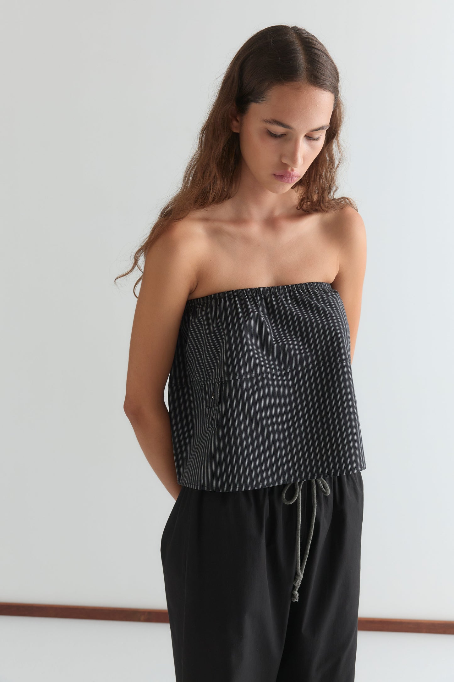 A woman with long brown hair stands against a white background, looking down. She wears a black strapless, loose top with thin white stripes and the Pocket Skirt - Mono Stripe, featuring an elasticated waistband and her arms behind her back.