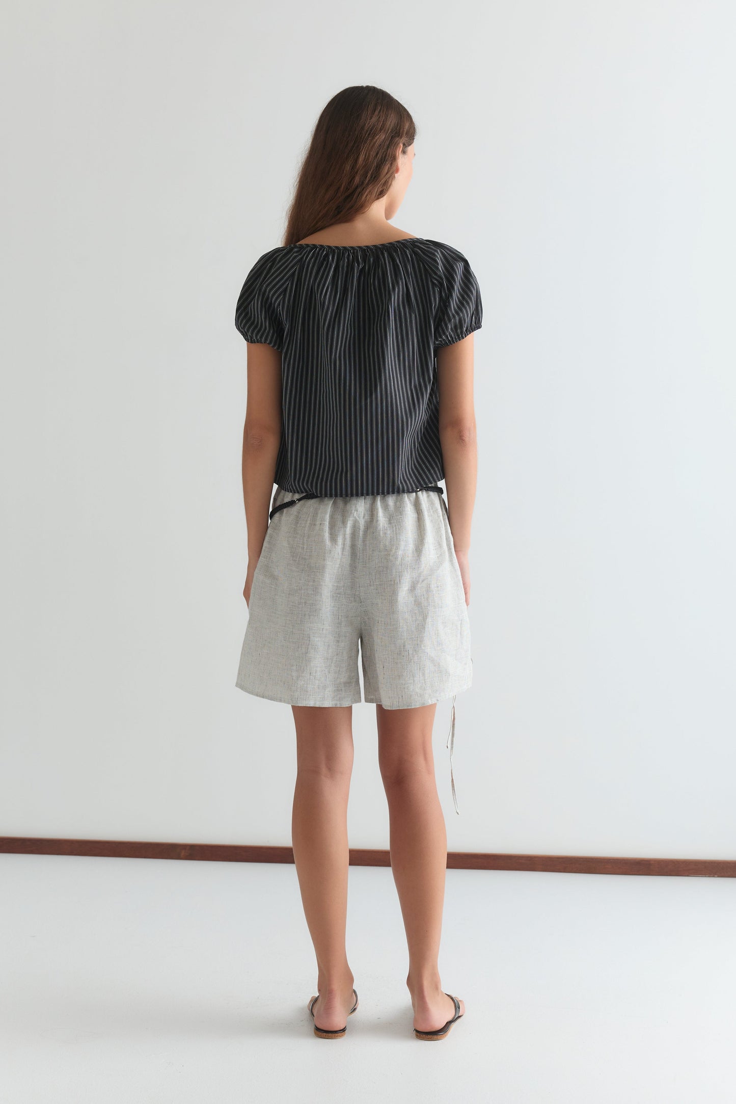 A woman with long brown hair, wearing the Poplin T Shirt - Mono Stripe by Deiji Studios and light gray shorts, stands with her back to the camera against a plain white background.