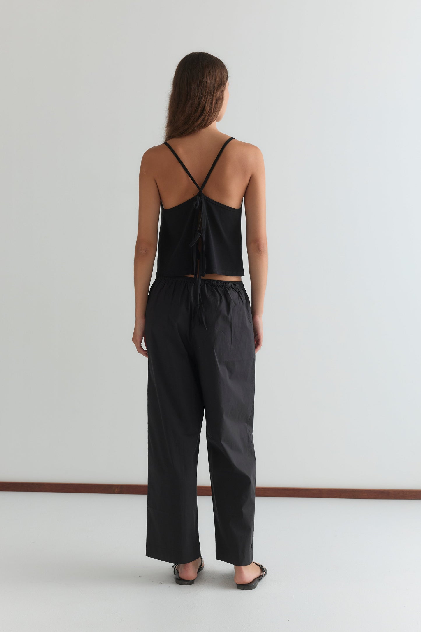 A woman stands with her back to the camera, wearing the Deiji Studios Drape Jersey Top in black, paired with loose black pants and black sandals. Her long brown hair falls down her back against a plain white wall.