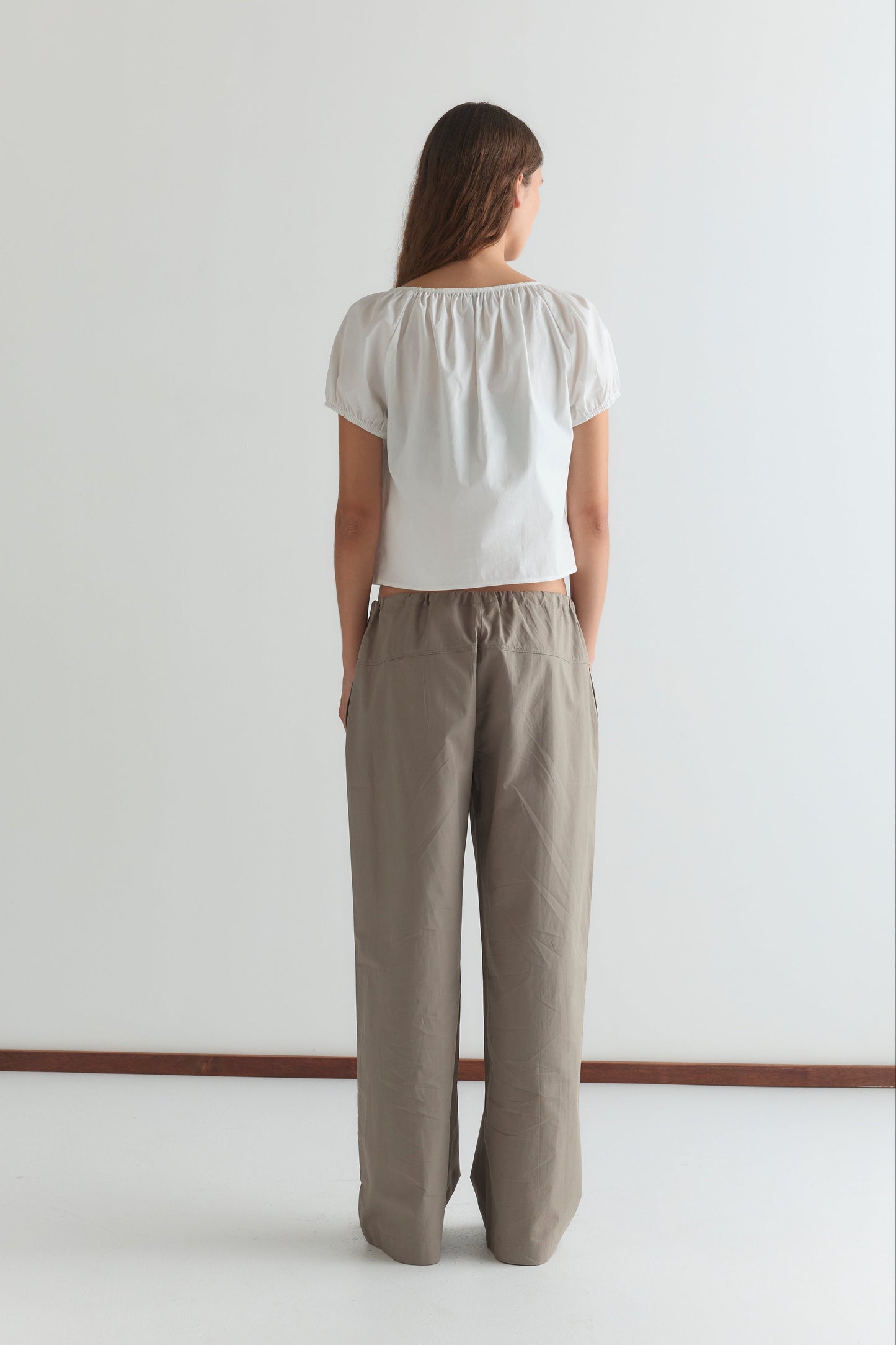 A person faces away from the camera, wearing the Poplin T Shirt - White by Deiji Studios with puffed sleeves and loose beige pants. The background is plain white with a brown strip along the floor.
