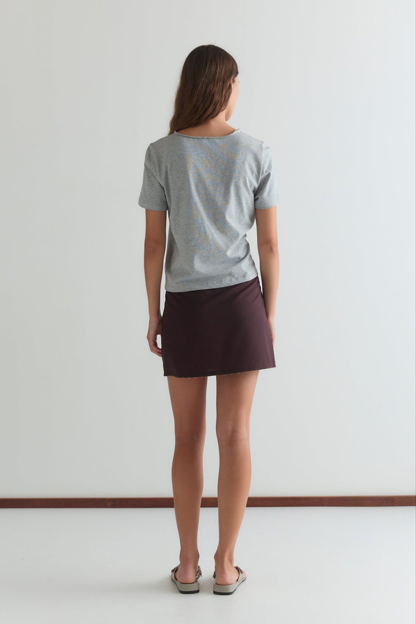 A woman with long brown hair stands facing away, wearing a gray t-shirt, the Eye Skirt in Burgundy Brown Stripe by Deiji Studios, and sandals. She is in front of a plain white wall and standing on a white floor.