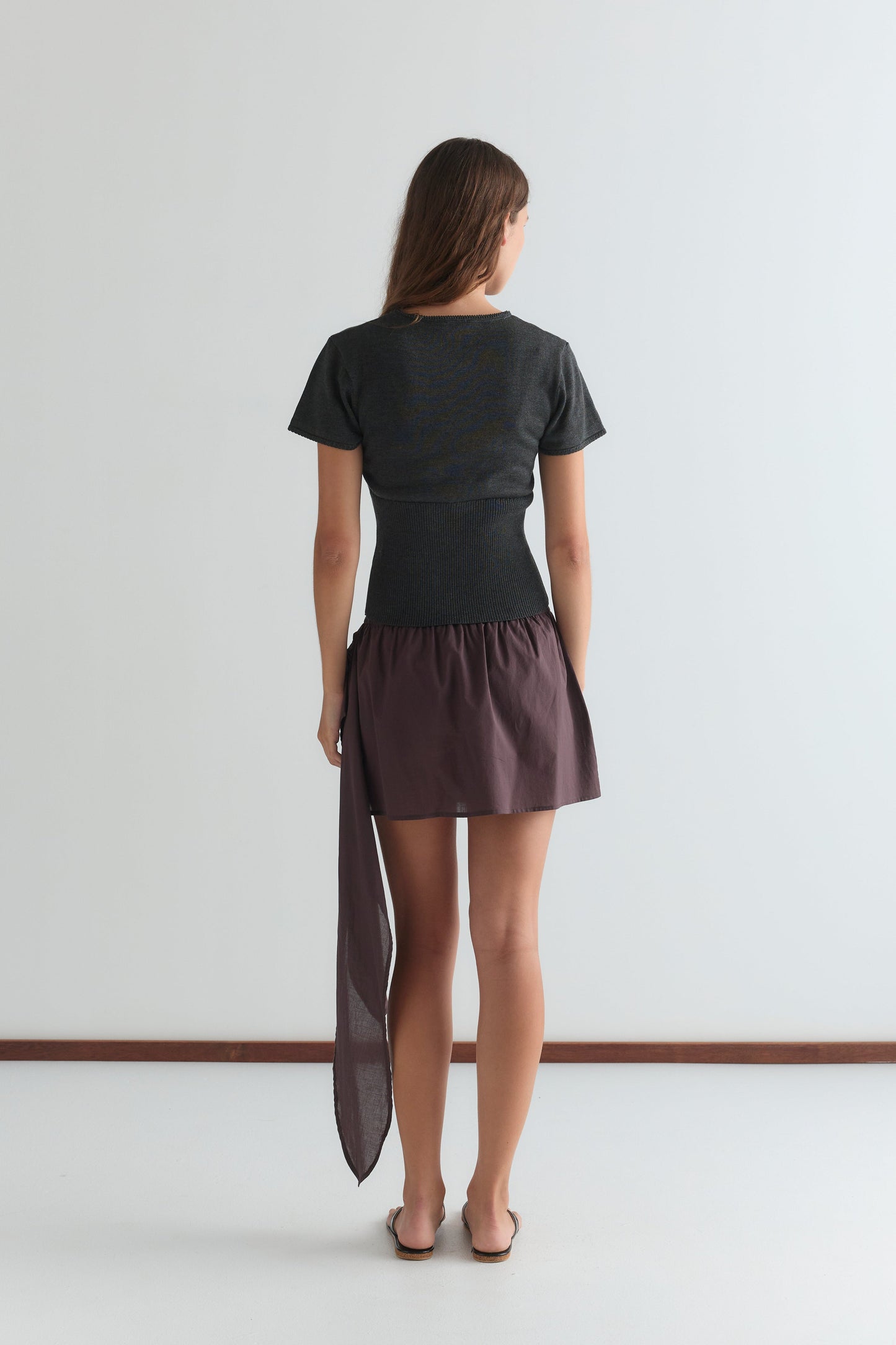 A woman, seen from behind against a plain white background, wears the Trim Knit Tee in dark grey with a short purple skirt featuring an asymmetrical long sheer fabric detail on one side.