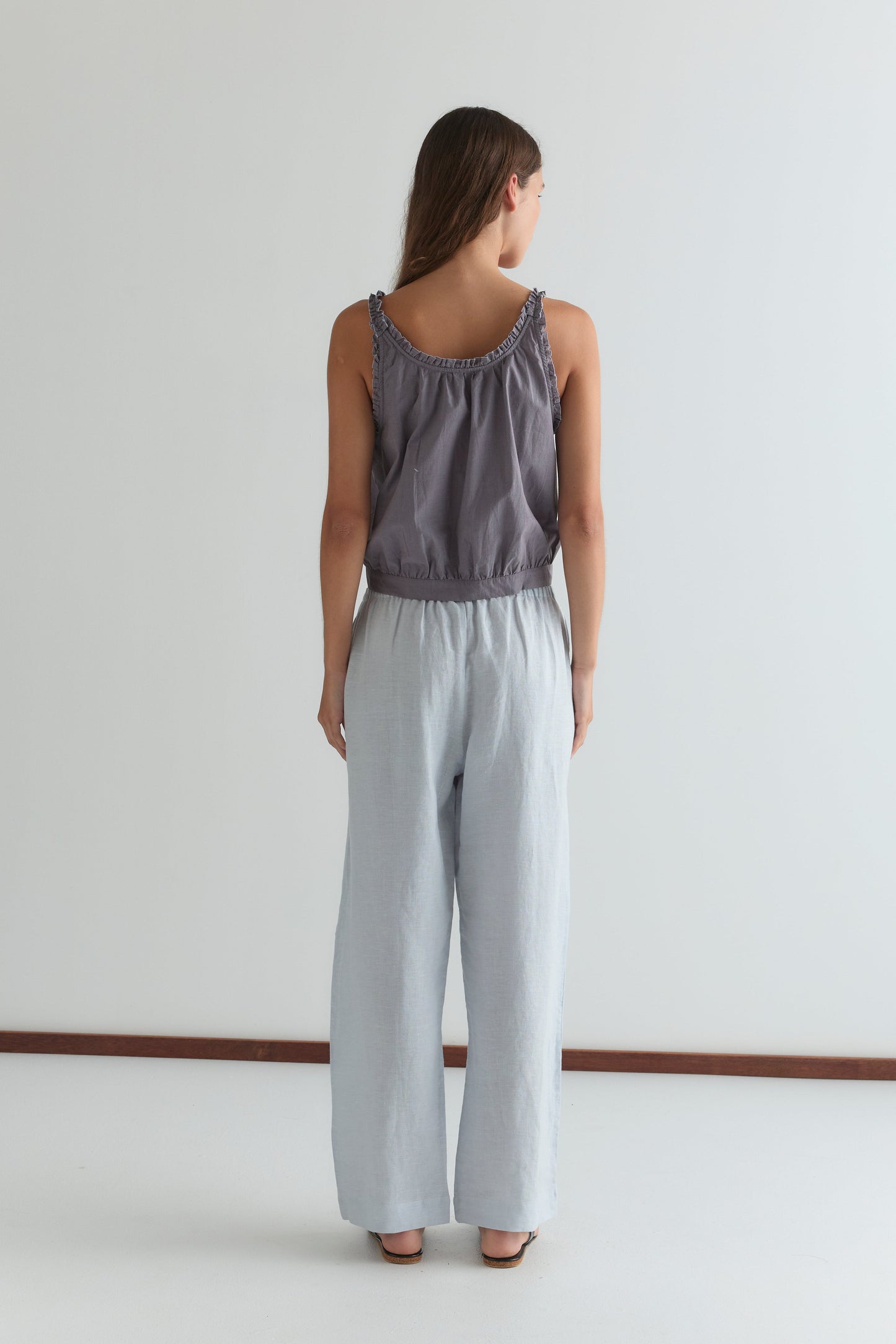 A woman faces away from the camera, wearing relaxed ankle-length Eyelet Pant in Soft Blue by Deiji Studios with a sleeveless, loose gray top, set against a plain white background.