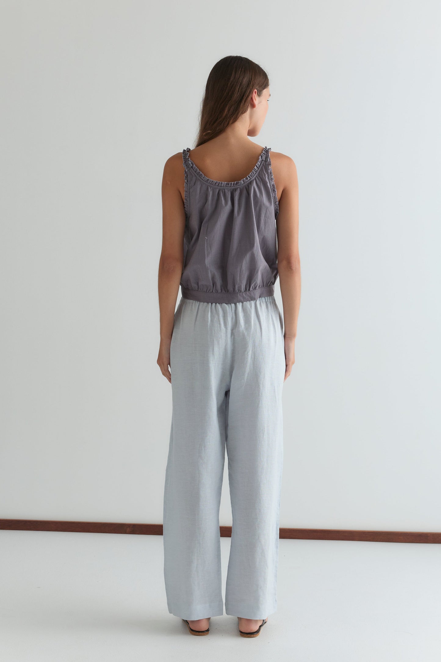 A woman stands facing away from the camera in the Overlie Top - Currant by Deiji Studios and light blue wide-leg pants, her hair down and straight against a plain white background.
