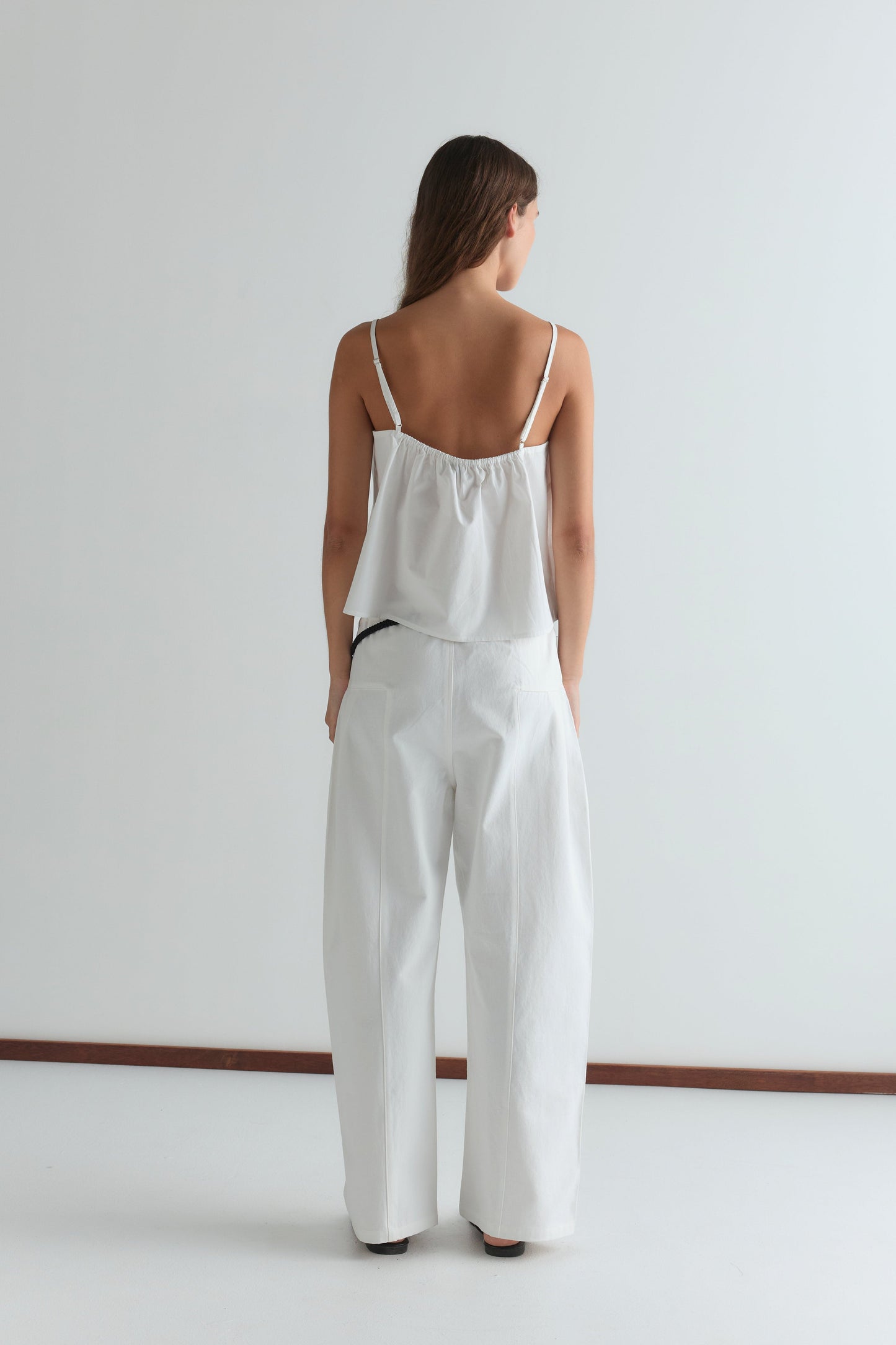 A person with long brown hair stands facing away, wearing the Double Layer Top - White from Deiji Studios and loose white pants in a minimalistic room with a white background.