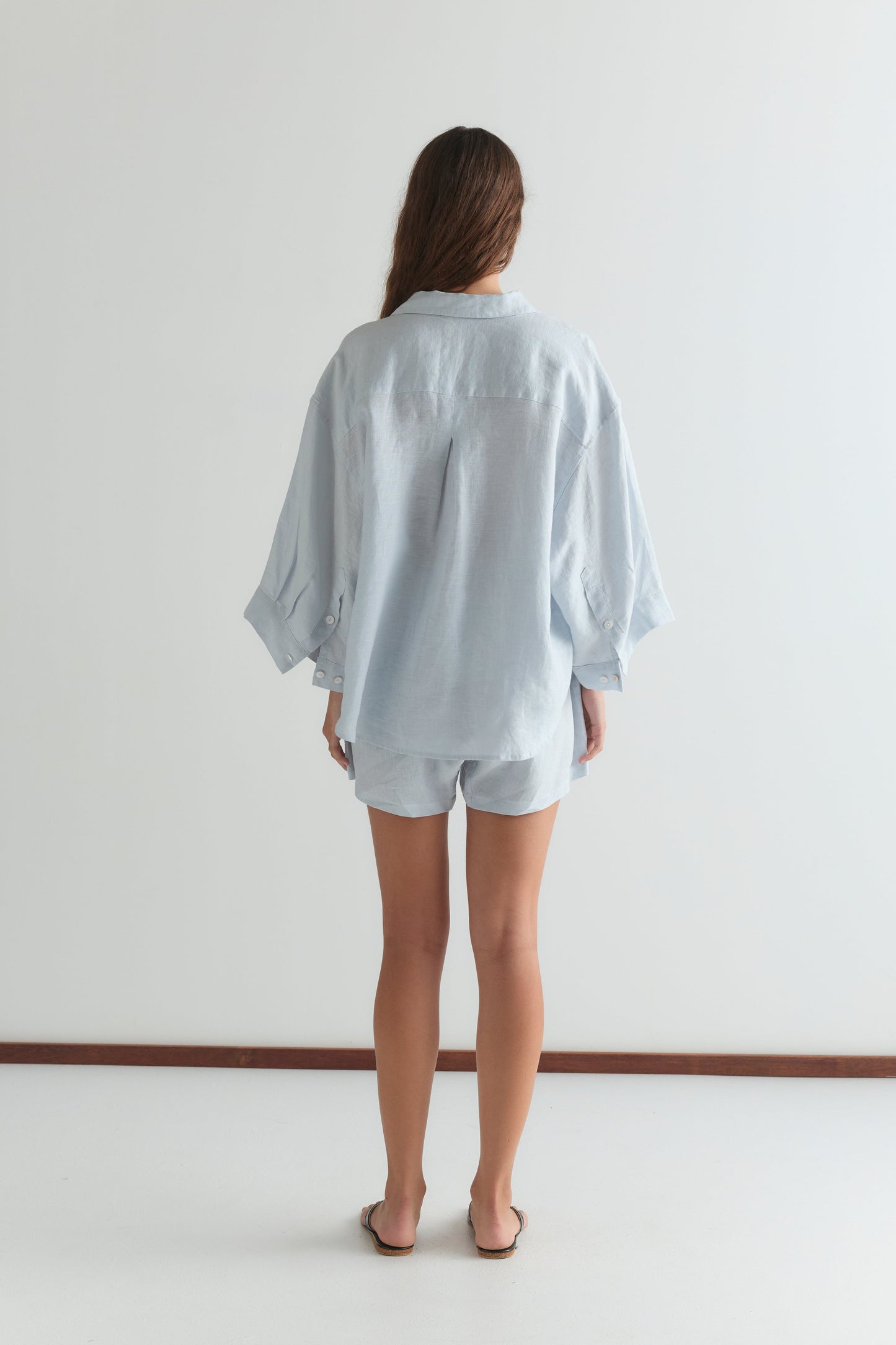 A woman, barefoot on a white floor against a plain white wall, wears the 03 Set - Soft Blue by Deiji Studios: an oversized linen shirt and matching boxer shorts.