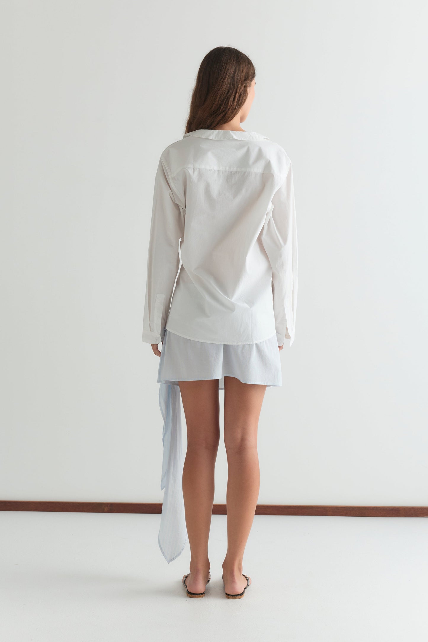 A woman, barefoot and facing away from the camera, wears the Left Shirt - White with a relaxed silhouette over a light blue skirt or shorts against a plain white background.