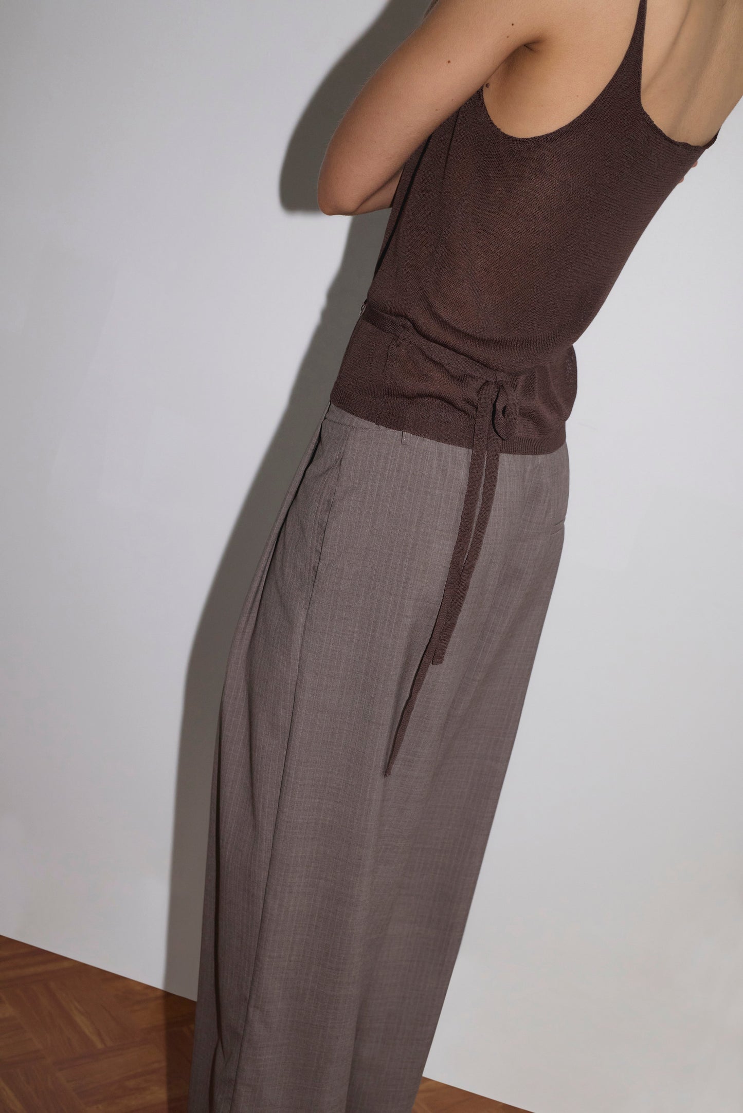 A person stands against a plain white wall, wearing the Square Knit Top - Brown by Deiji Studios with a waist tie and loose gray trousers. The person's head and part of their arms are out of the frame.