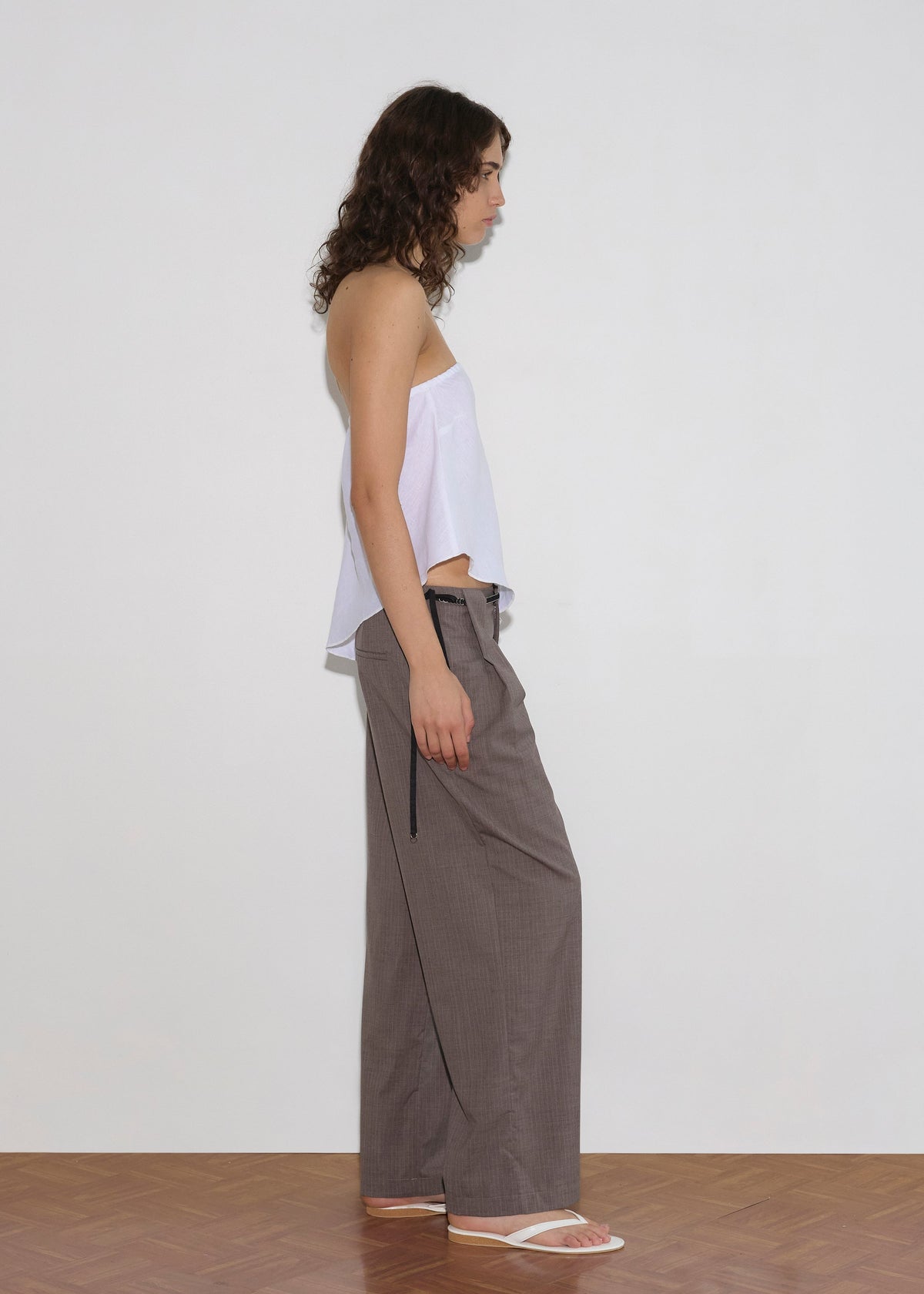A person with medium-length curly hair stands in profile against a plain white wall, wearing the Line Top - White from Deiji Studios, loose gray trousers with a black drawstring and asymmetric hem, and white sandals on a wooden floor.