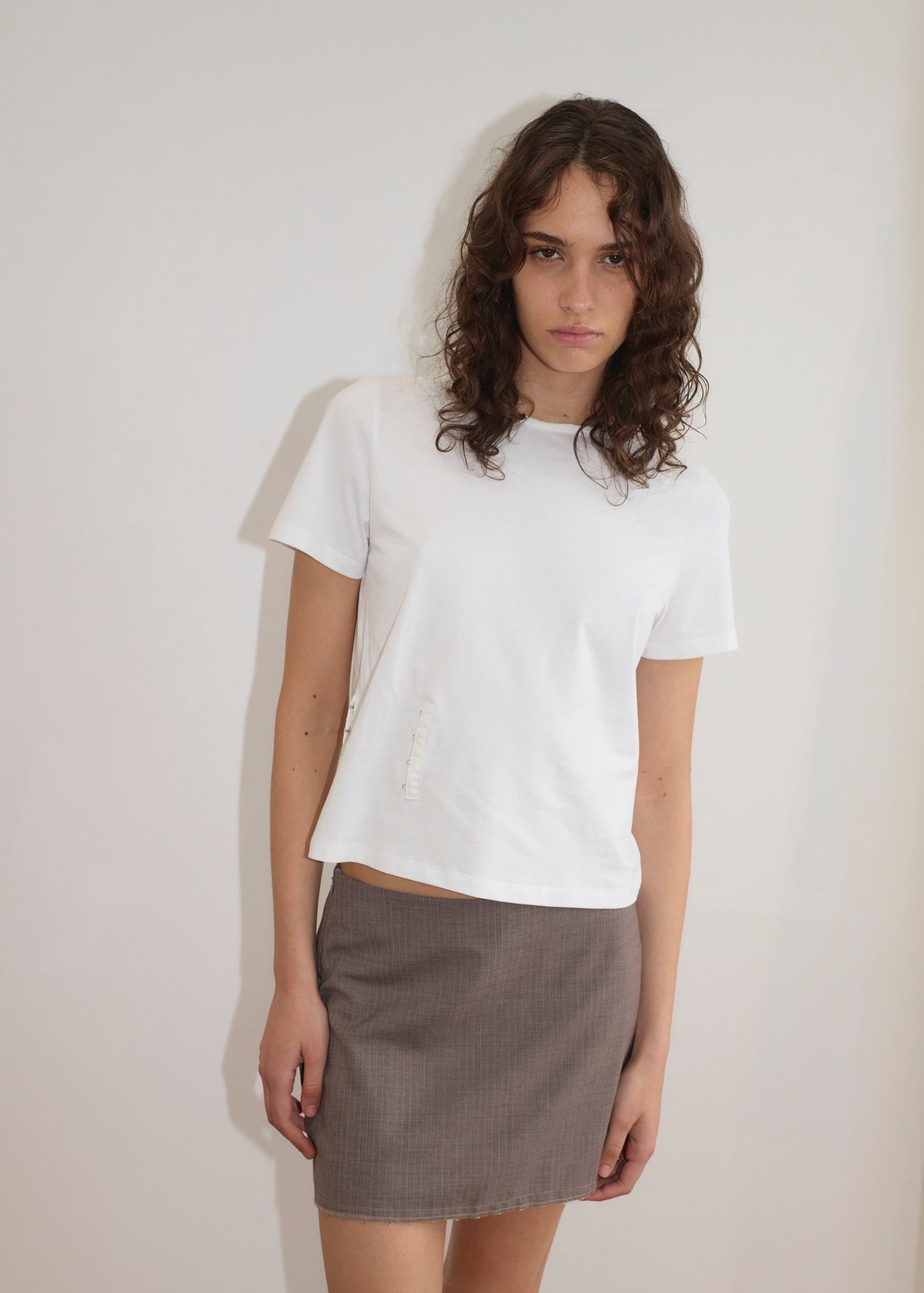 A young woman with wavy brown hair, wearing a plain white t-shirt and the Eye Skirt in Grey Pink Stripe from Deiji Studios, stands against a plain white wall, looking at the camera with a neutral expression.