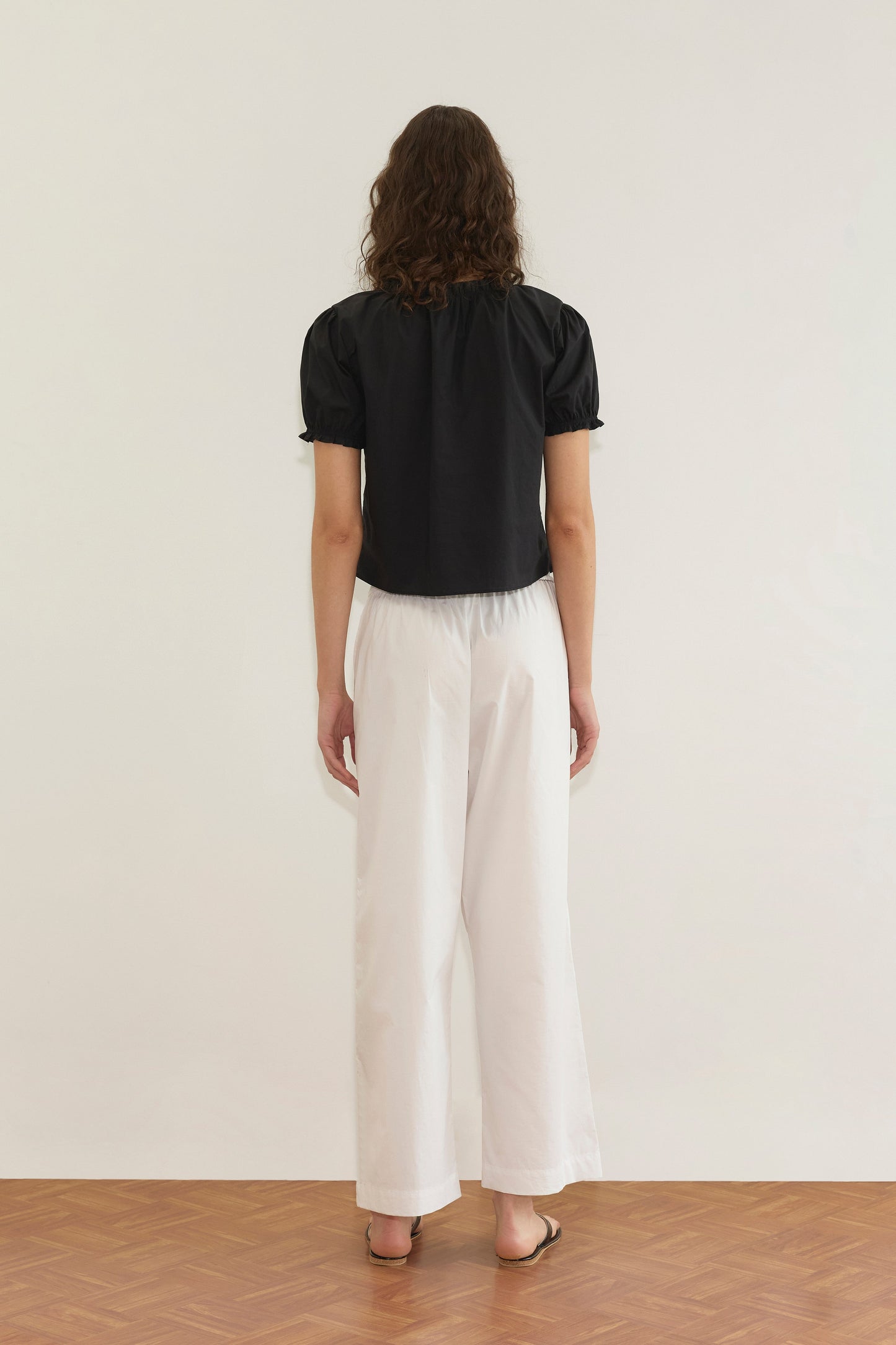 A person with wavy brown hair stands facing away, wearing the Cross Frill Top - Black with a cropped relaxed silhouette, paired with loose white pants and brown sandals on a wooden floor against a plain white wall.