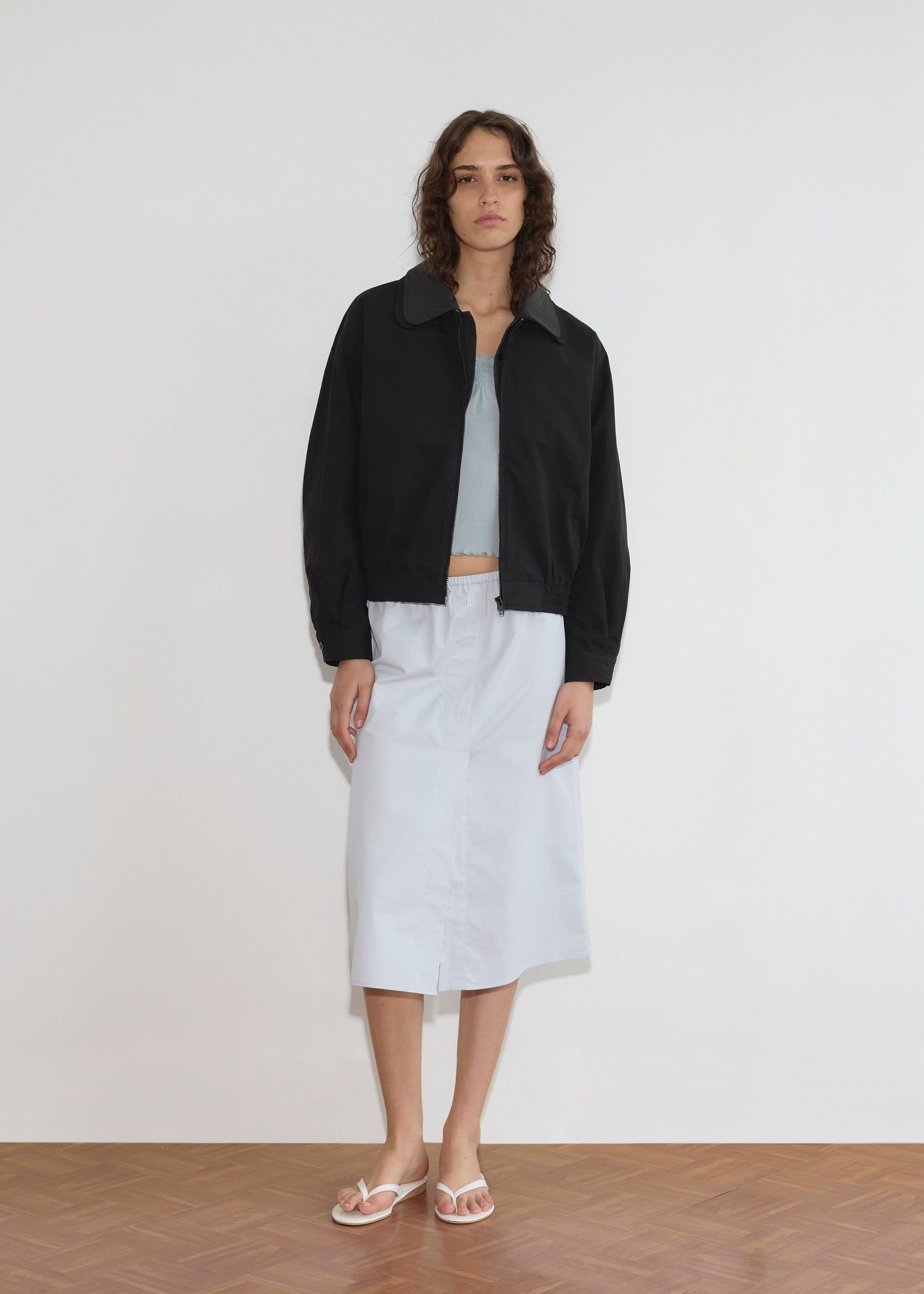 A person with wavy hair stands indoors against a white wall, wearing the Collared Zip Jacket - Black Contrast, a light blue top, white midi skirt, and white flip-flops on a wooden floor.