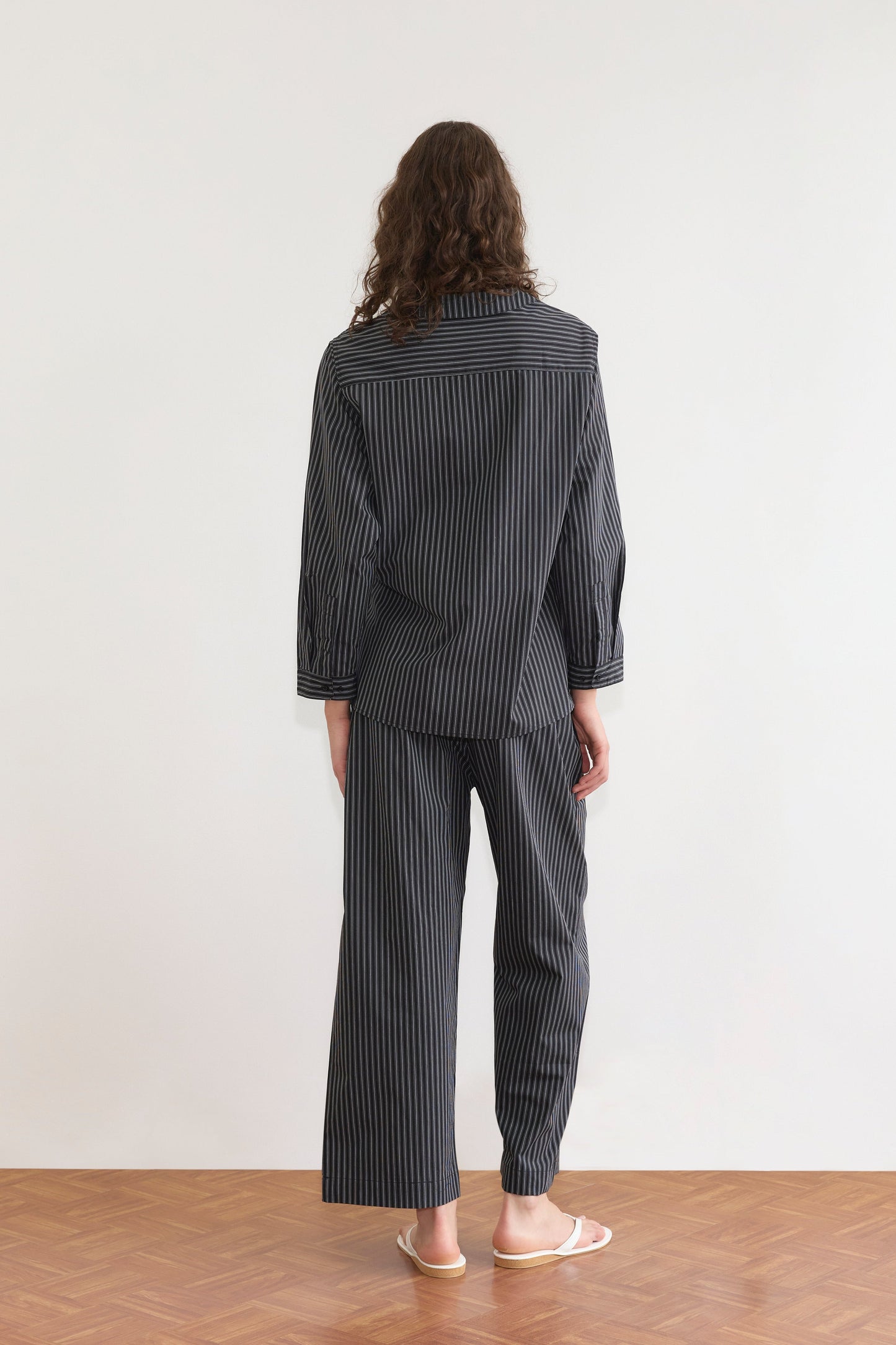 A person with long curly hair, seen from behind, wears the Left Set - Mono Stripe by Deiji Studios and white flip-flops, standing on a wooden floor against a plain light-colored wall.