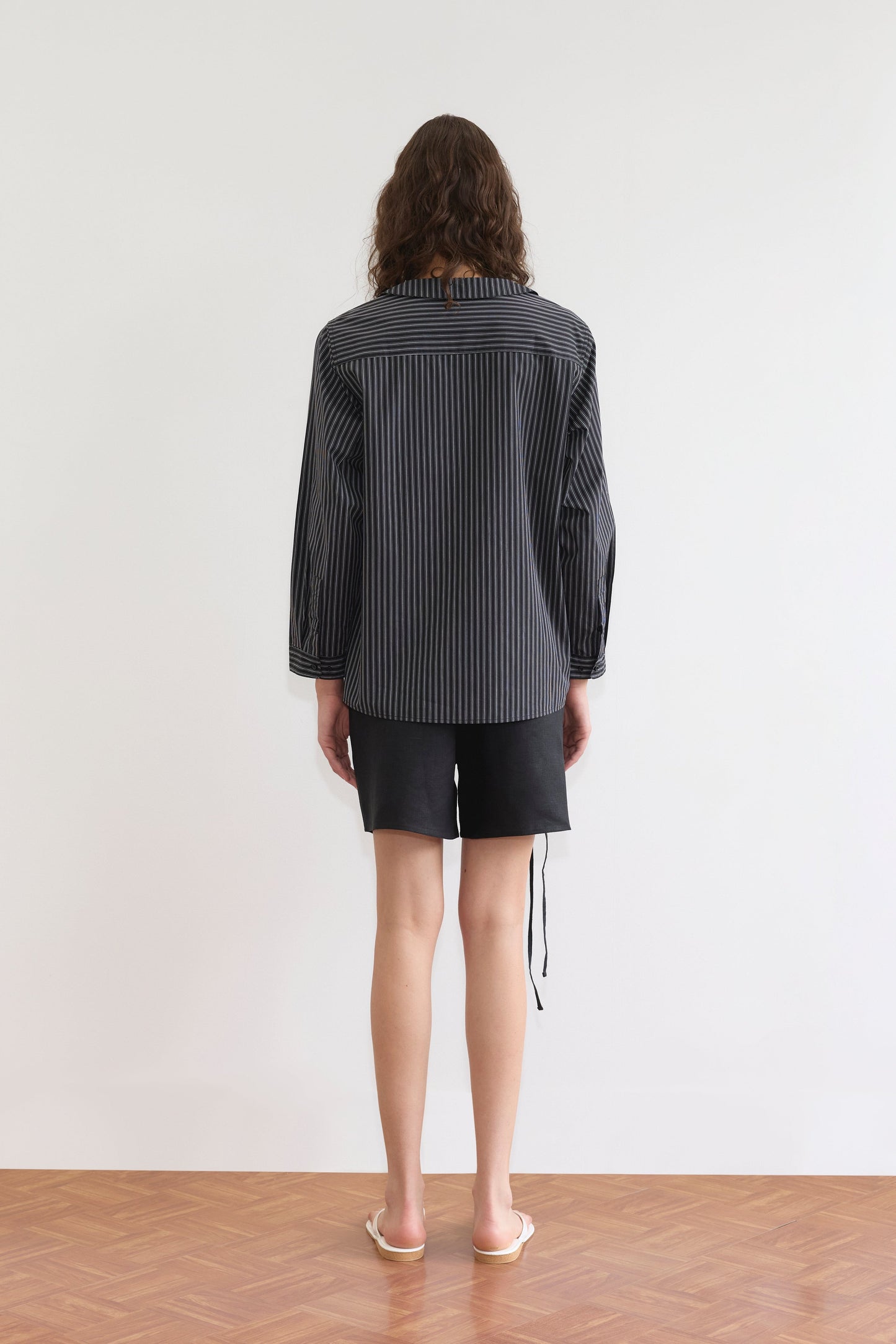 A person with shoulder-length brown hair faces a white wall, wearing the Deiji Studios Left Shirt - Mono Stripe, black shorts, and beige flip-flops on parquet flooring for a relaxed silhouette.