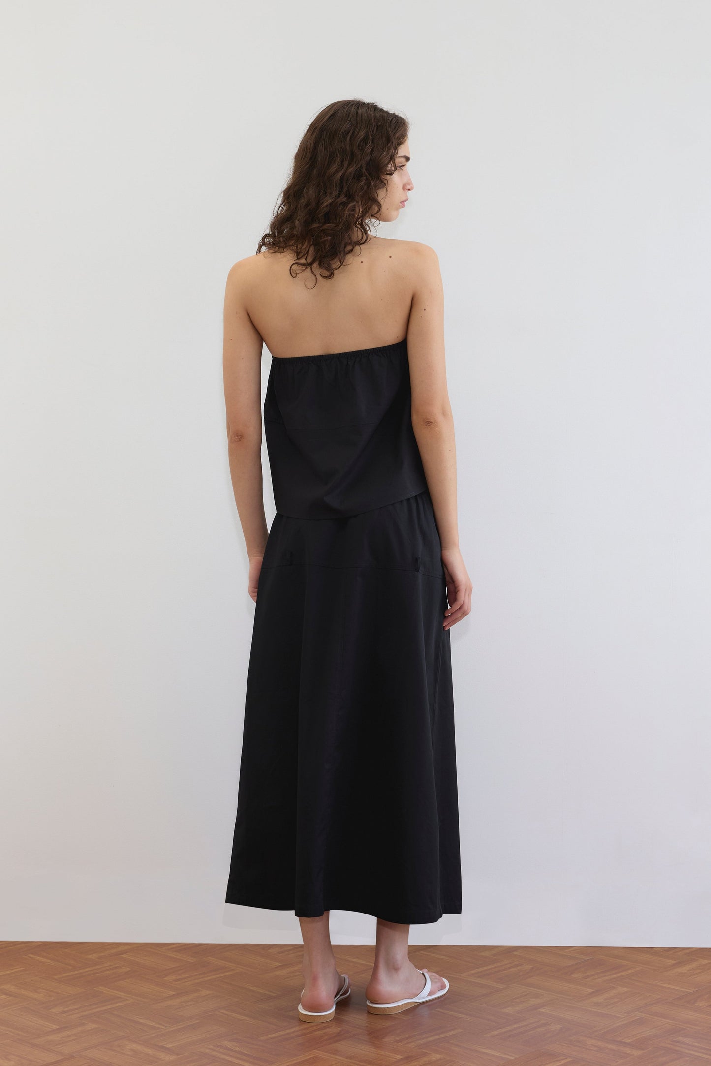 A woman with curly hair, barefoot on a wooden floor, stands facing away in a strapless black top and the Loop Pocket Skirt - Black by Deiji Studios, set against a plain white wall.