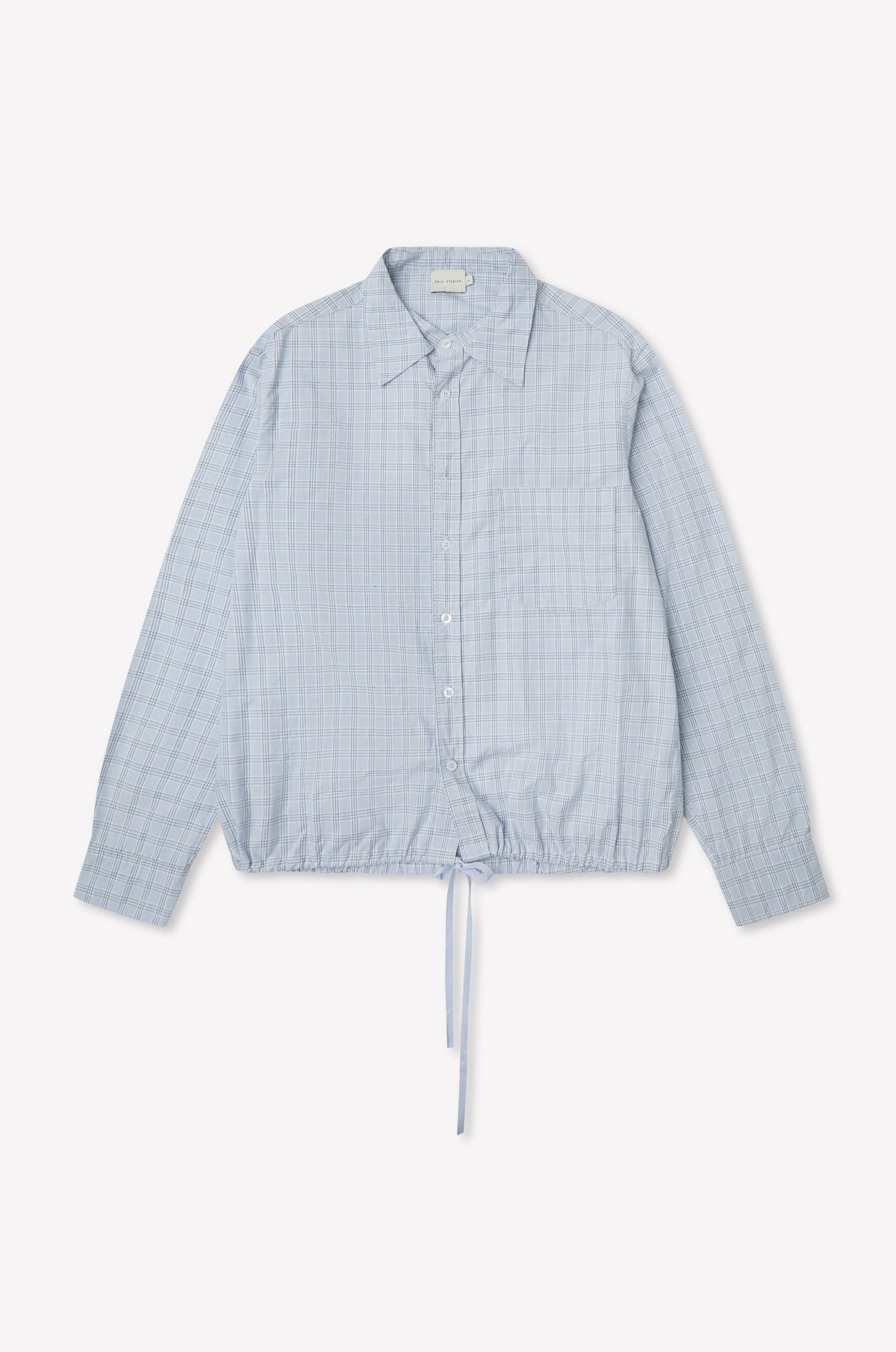 The Stand Shirt - Powder Check is a light blue oversized cotton shirt with long sleeves, subtle checkered pattern, chest pocket, classic collar, and drawstring hem. Shown on a plain white background, it channels deiji studios' relaxed style.