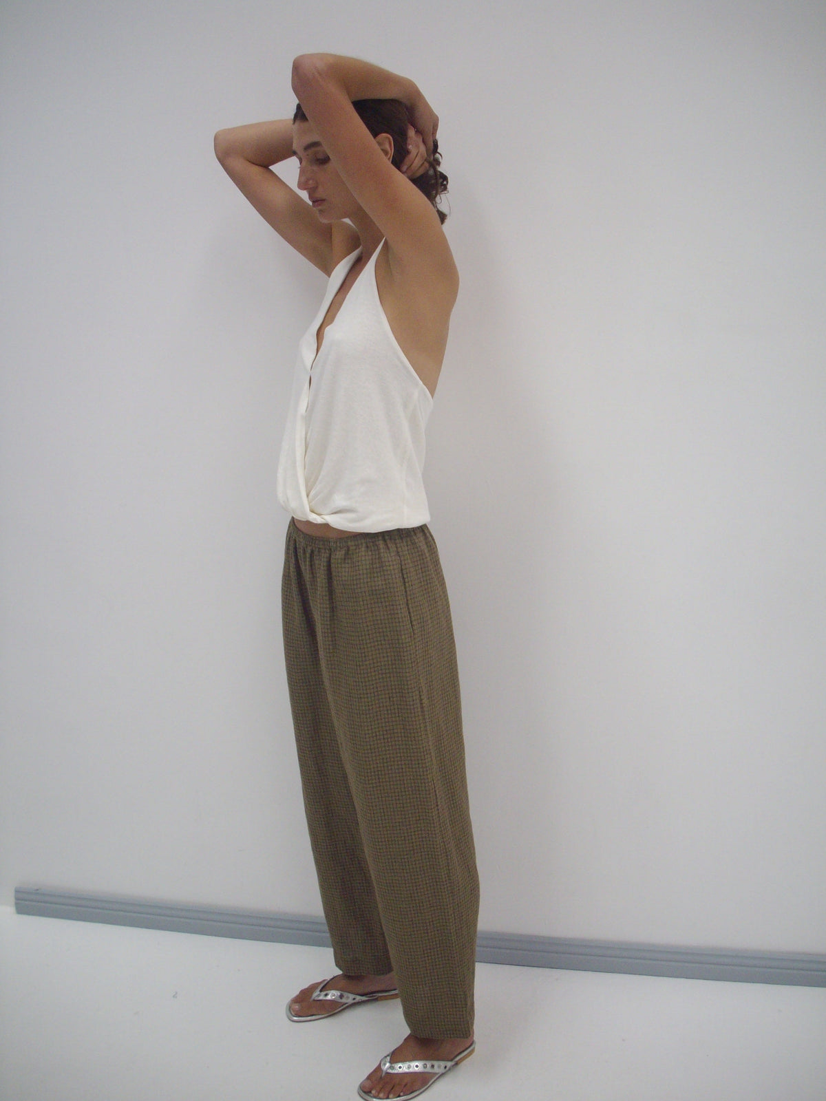 A person stands sideways by a white wall, wearing a loose white sleeveless top, Linen Ease Trouser - Pine Check, and sandals. Their hands are raised behind their head, giving off a relaxed vibe.