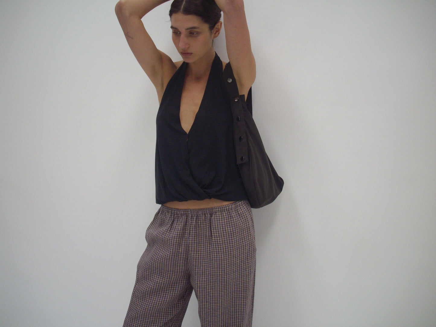 Wearing a sleeveless black V-neck top and Deiji Studios' linen ease trouser in russet check, a person with dark hair tied back stands against a white wall, holding a dark bag on one shoulder.