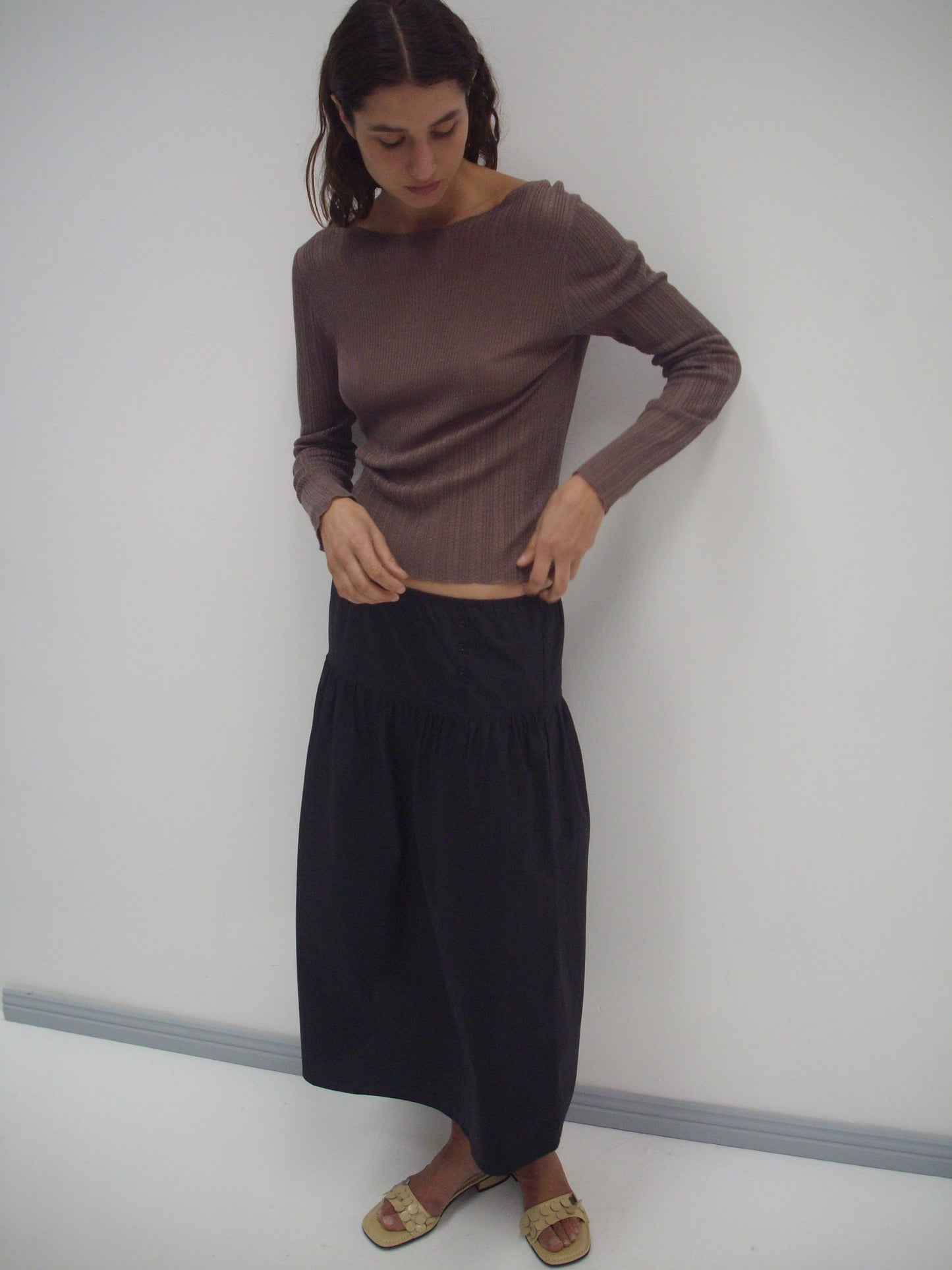 A woman with shoulder-length hair stands against a white wall, wearing a fitted brown long-sleeve top, the Deiji Studios Button Skirt in black, and beige sandals while looking down.