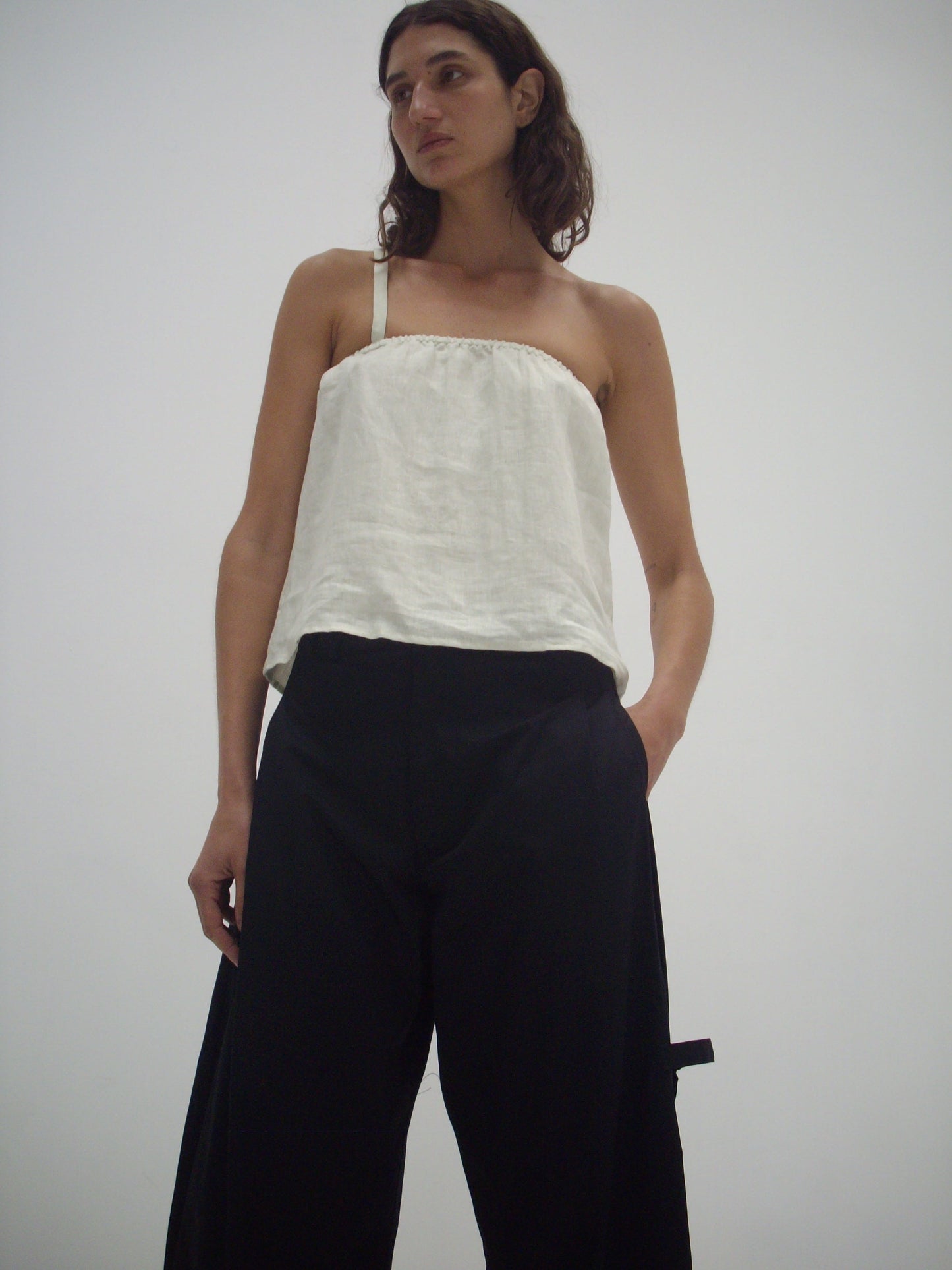 A person with long brown hair wears a loose white strapless top and the Tab Pant - Black from Deiji Studios, standing against a plain white background with hands in their pockets, looking slightly to the side.