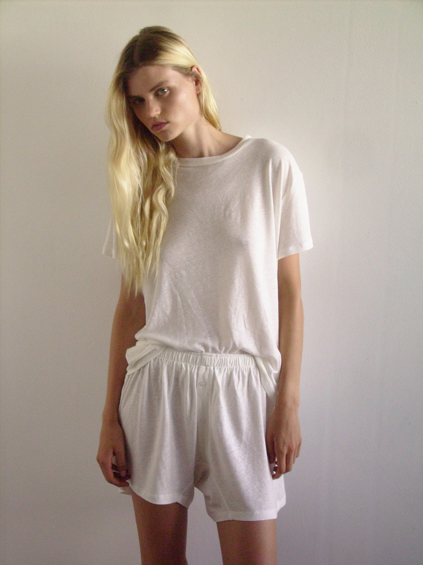 A person with long blonde hair stands against a white wall, wearing a loose white loungewear T-shirt and the soft short - ecru, looking slightly to the side with a neutral expression.