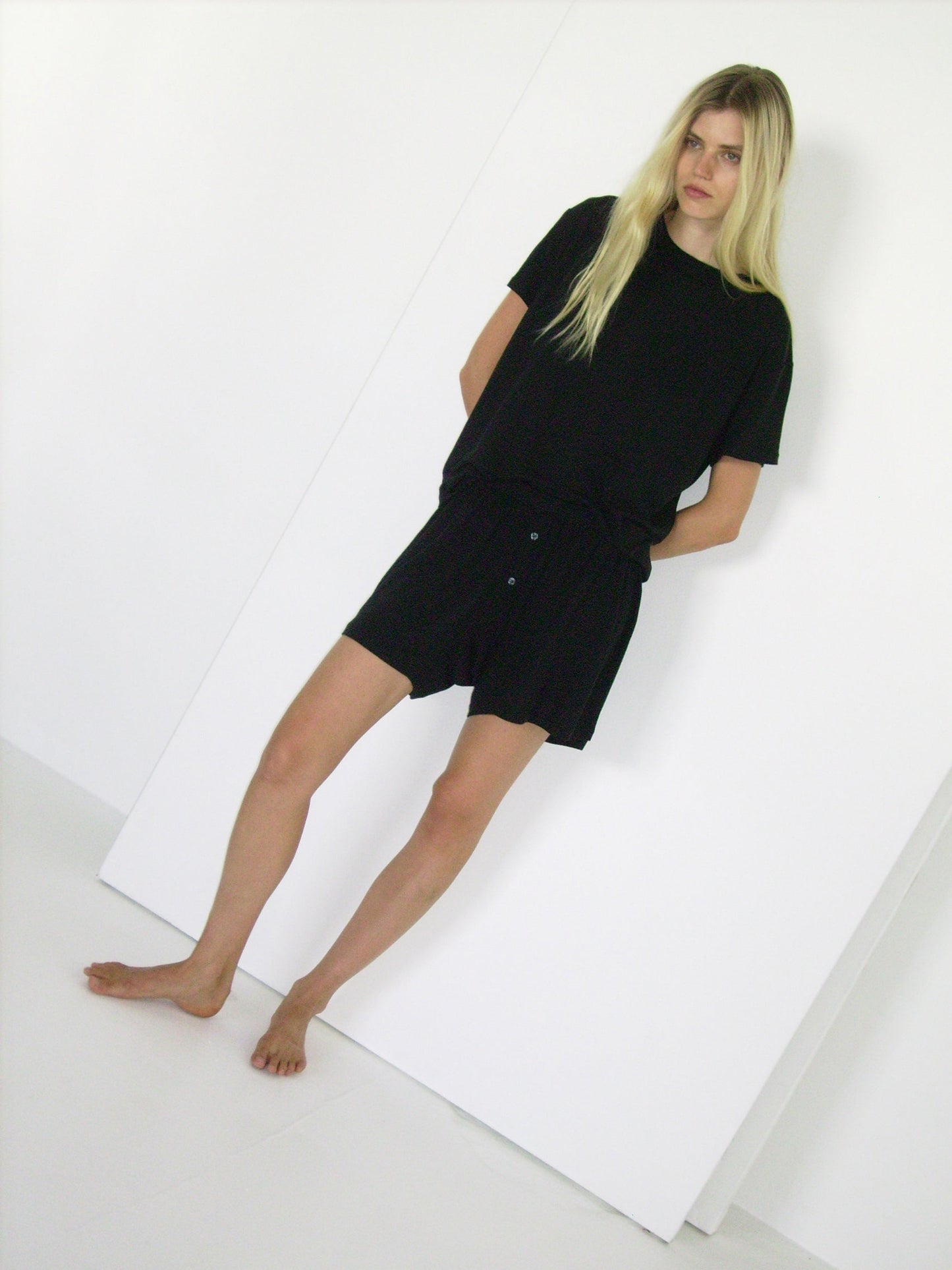 A woman with long blonde hair stands barefoot against a white background, wearing the Deiji Studios soft t shirt - black and matching black shorts, with her hands in her pockets.