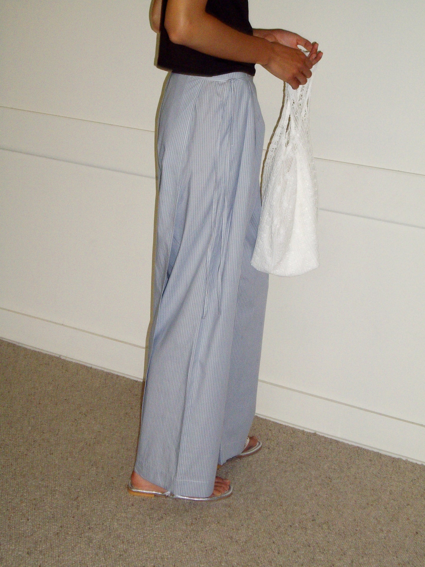 Wearing the Double Layer Pant - Sky Stripe with a black top and sandals, a person holds a white textured bag while standing indoors against a plain wall and carpeted floor.