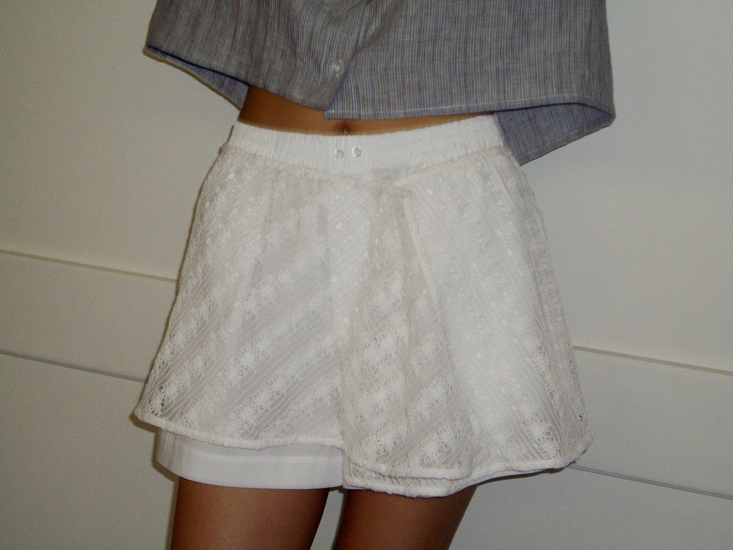 A person stands against a light wall wearing a gray pinstriped collared shirt and the Thread Button Belt - Off White, featuring white, lace-patterned organic cotton shorts. Focus is on the unique texture of the shorts.