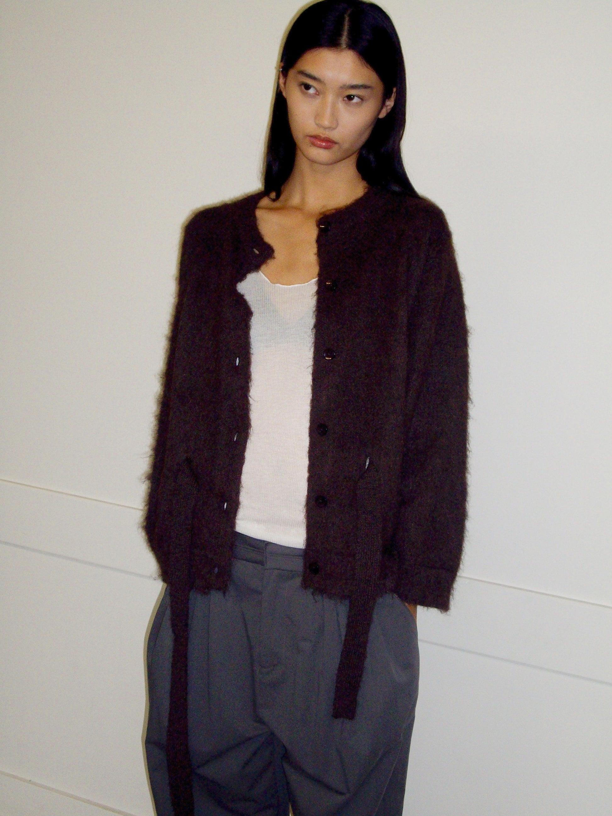 A person with long dark hair stands against a white wall, wearing the 0 Belted Cardi - Brown over a white tank top and gray pants, looking slightly to the side with a neutral expression.